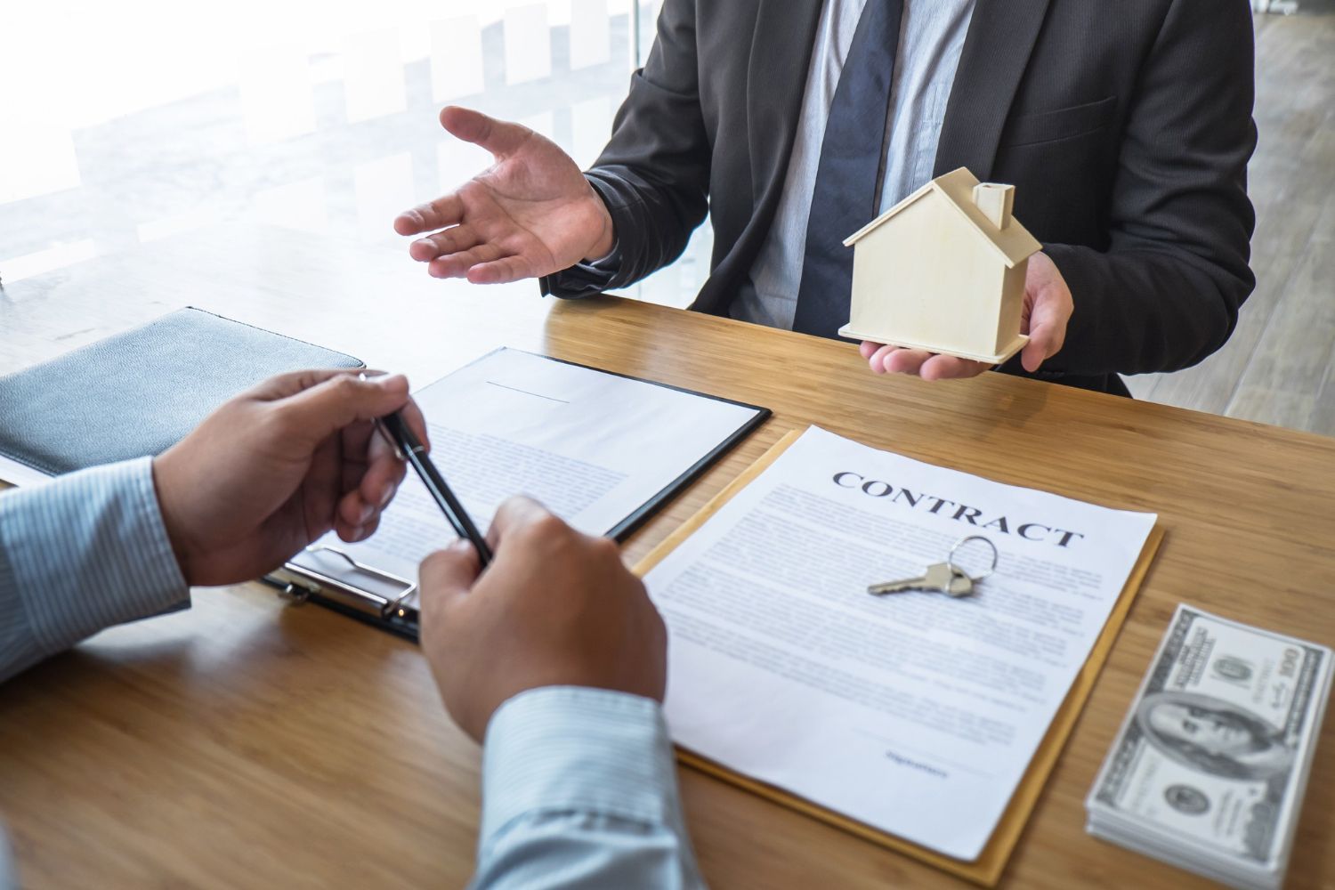 A person holding a model house sits across from someone with a pen and contract on a desk with a stack of cash.