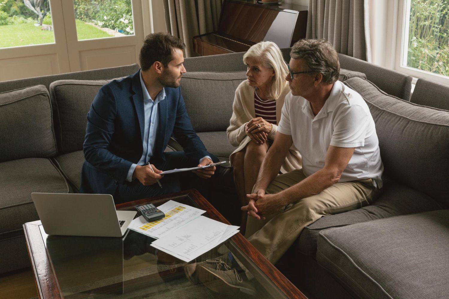 A professional advisor sits with a couple on a couch, reviewing documents on a coffee table with a laptop.
