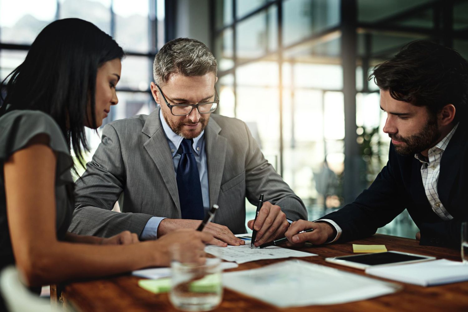 A professional meeting around a wooden table in a well-lit office, with three individuals reviewing documents.