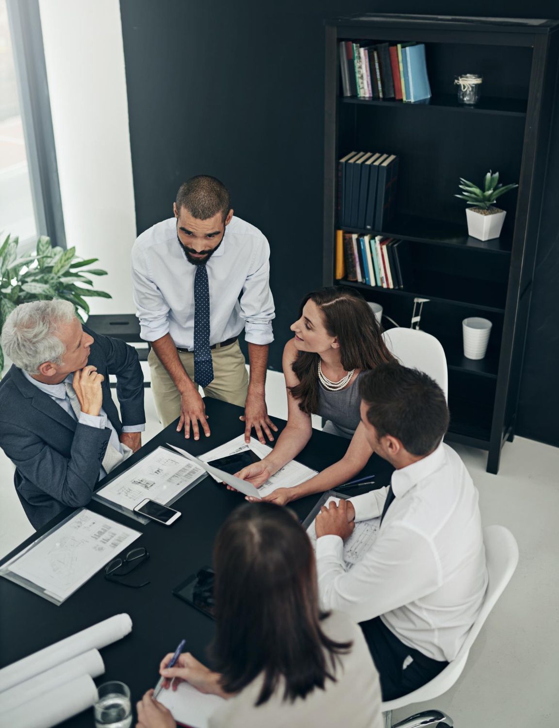 A diverse group of colleagues collaborate around a boardroom table with documents, looking toward a standing presenter.