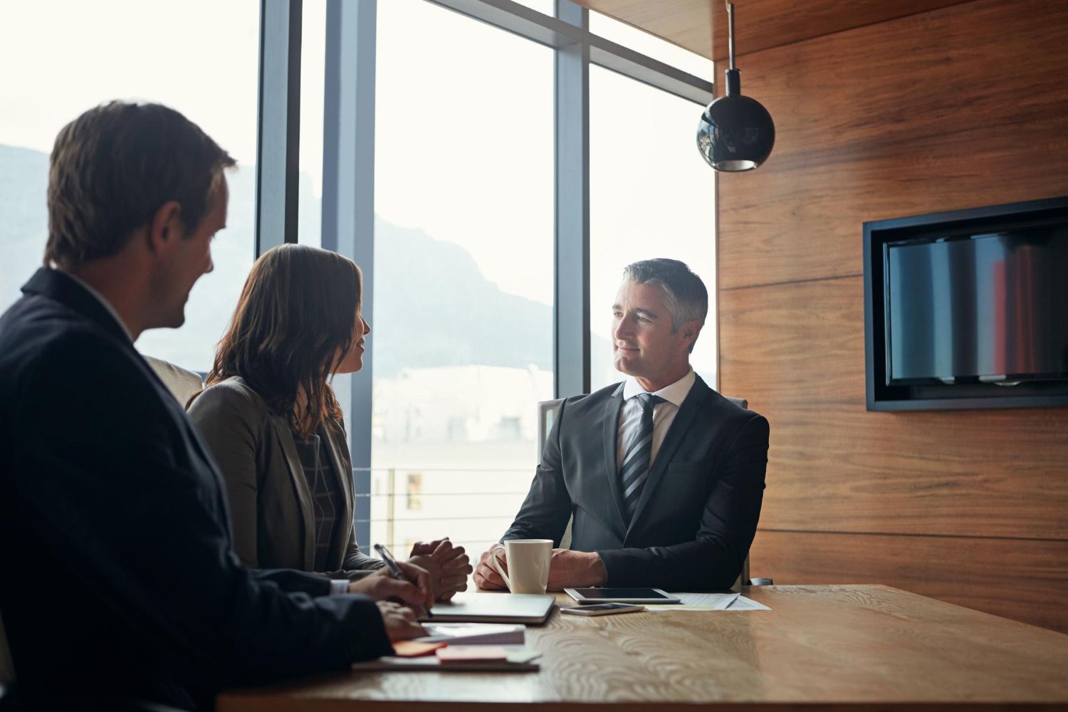 Three professionals in business attire discuss work at a wooden table in a bright, modern office with large windows.