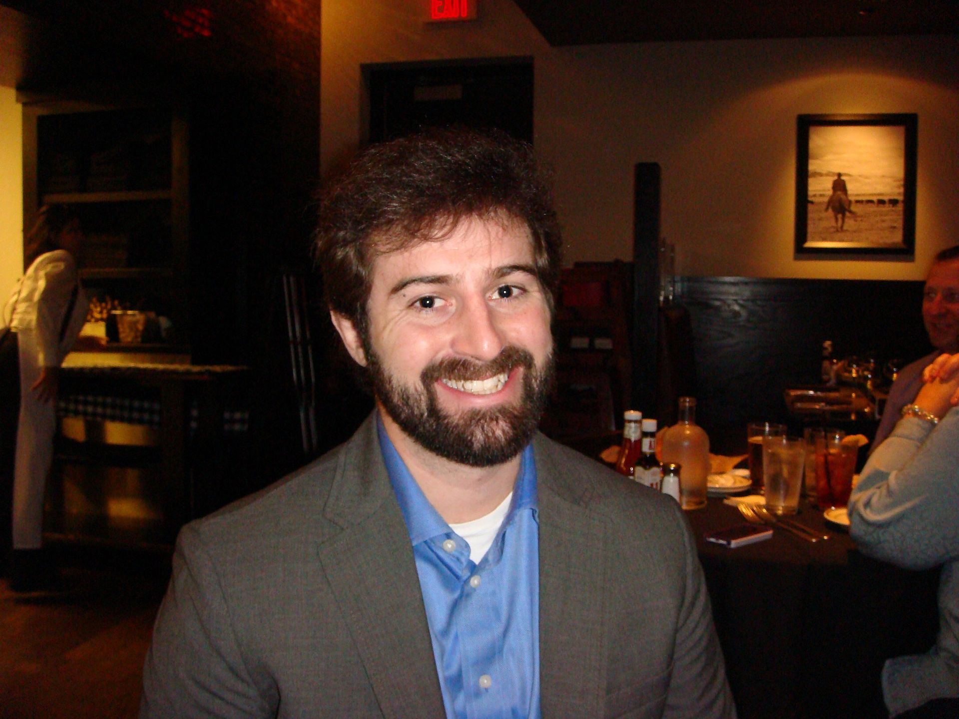 A smiling person with a beard, wearing a blue shirt and grey blazer, sitting in a dimly lit restaurant.