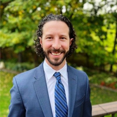 A smiling person with curly brown hair, wearing a blue suit, white shirt, and striped tie, outdoors against green foliage.