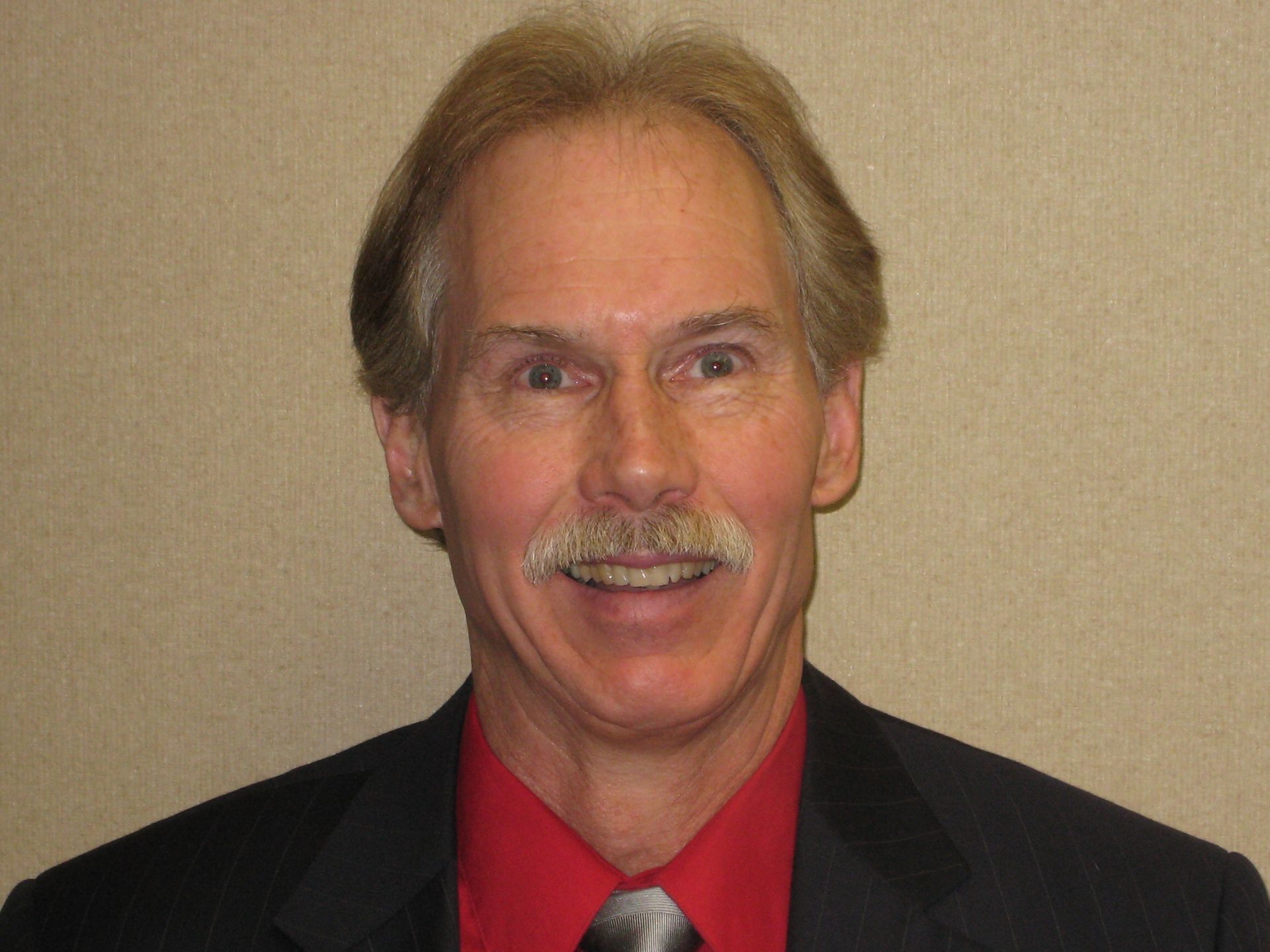 Head-and-shoulders portrait of a person with a mustache, wearing a dark suit, red shirt, and patterned necktie.
