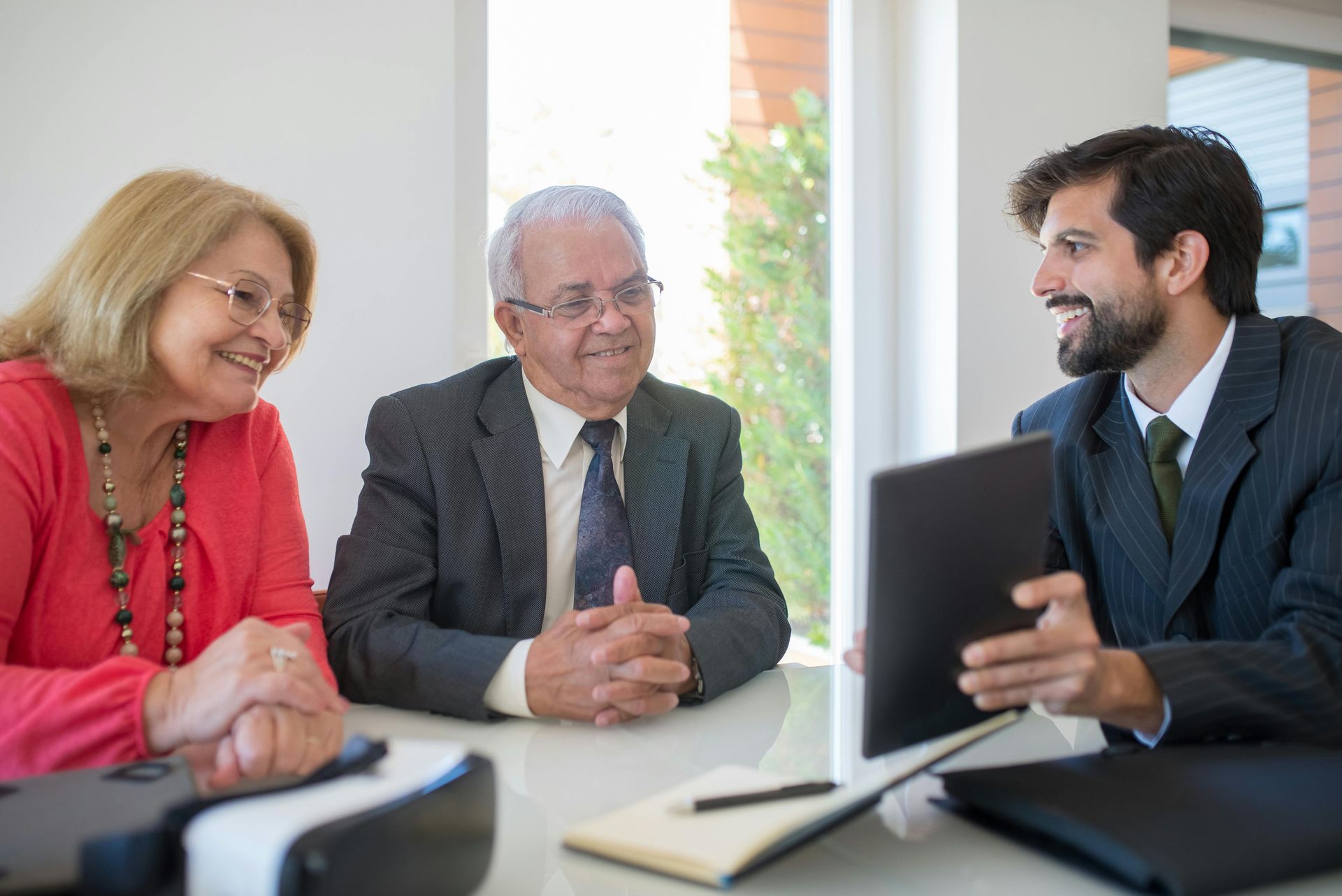 A man in a suit showing a tablet to two people at a desk in a bright office.