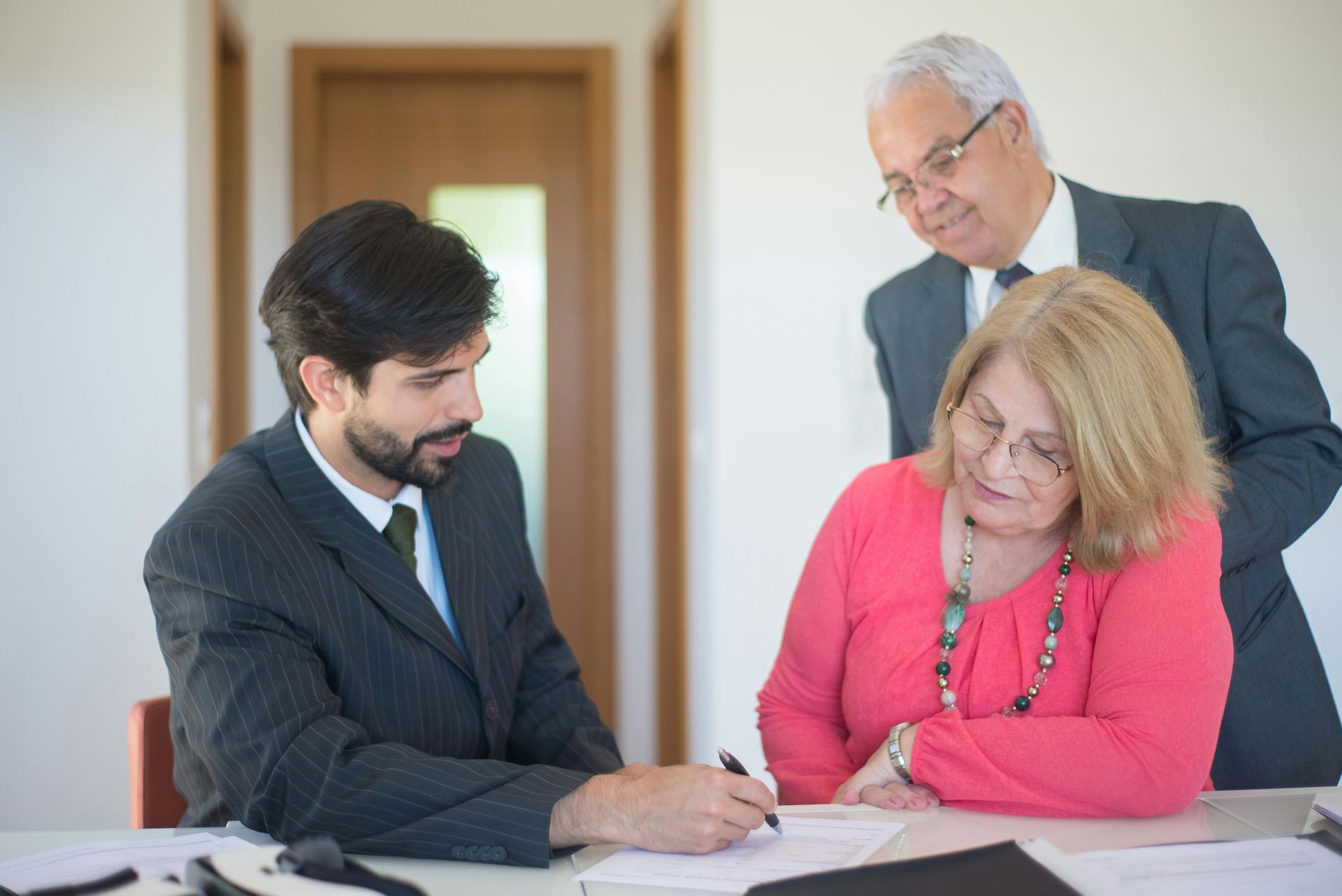 A man in a suit signs a document as a woman in a pink top and an older man in a suit watch him in an office.