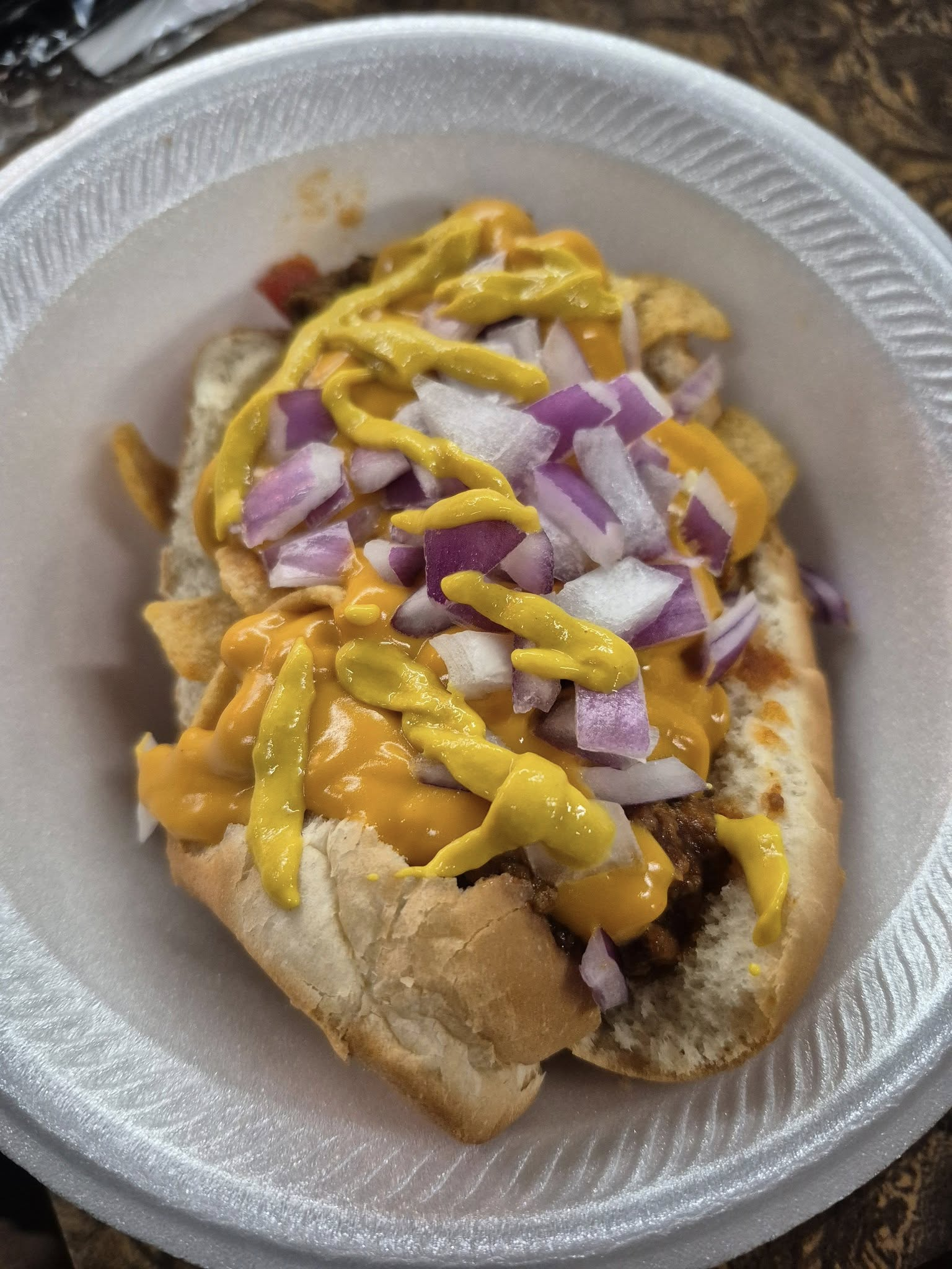 Hot dog on a bun, topped with cheese sauce, onions, and mustard, in a paper bowl.