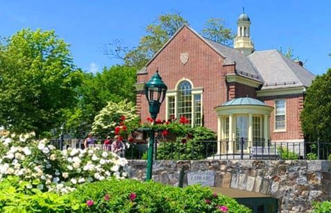 A brick building with a clock tower is surrounded by flowers and trees.