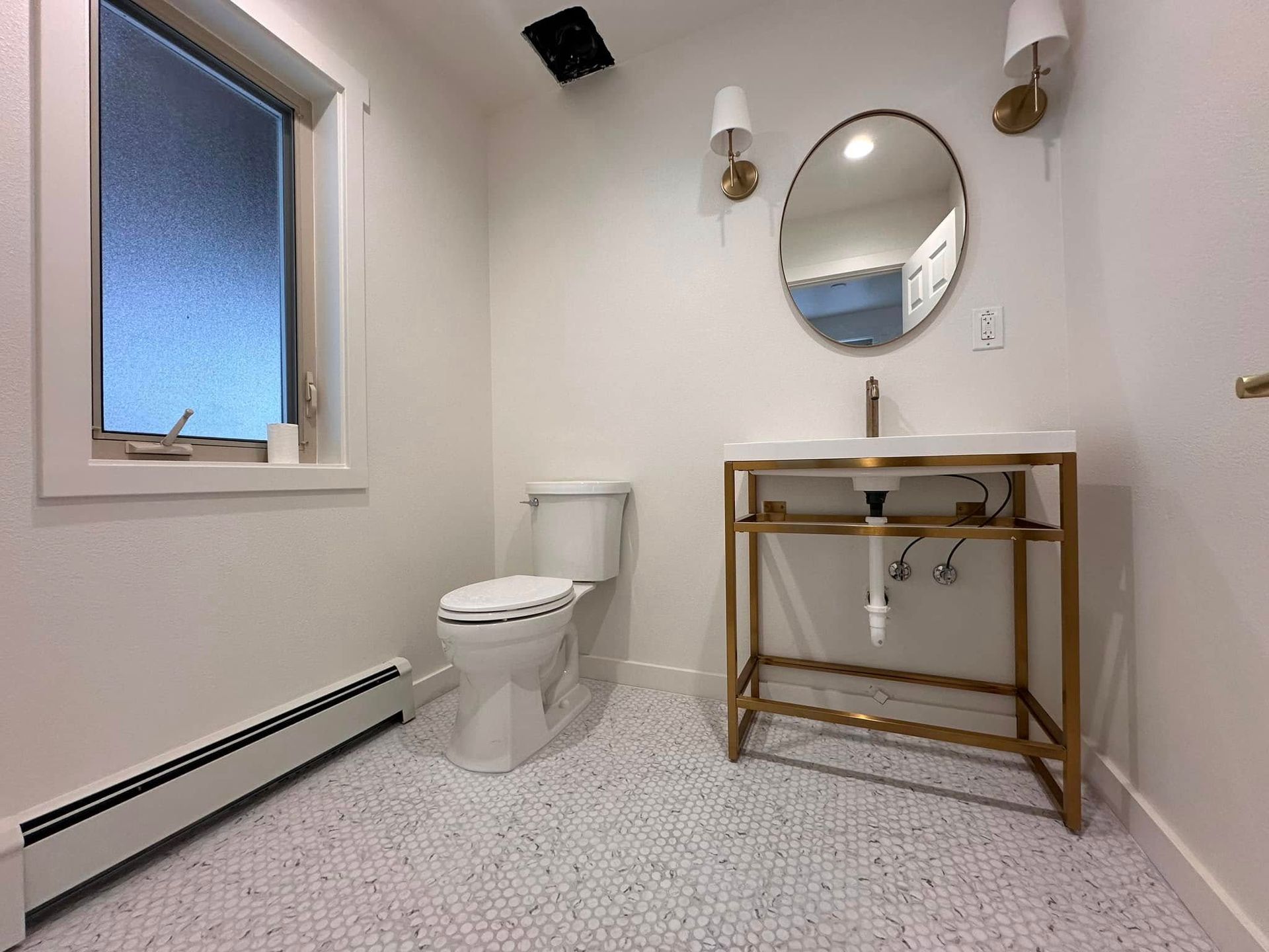 White bathroom with toilet, sink, and oval mirror; gold fixtures, white and pebble floor tile.