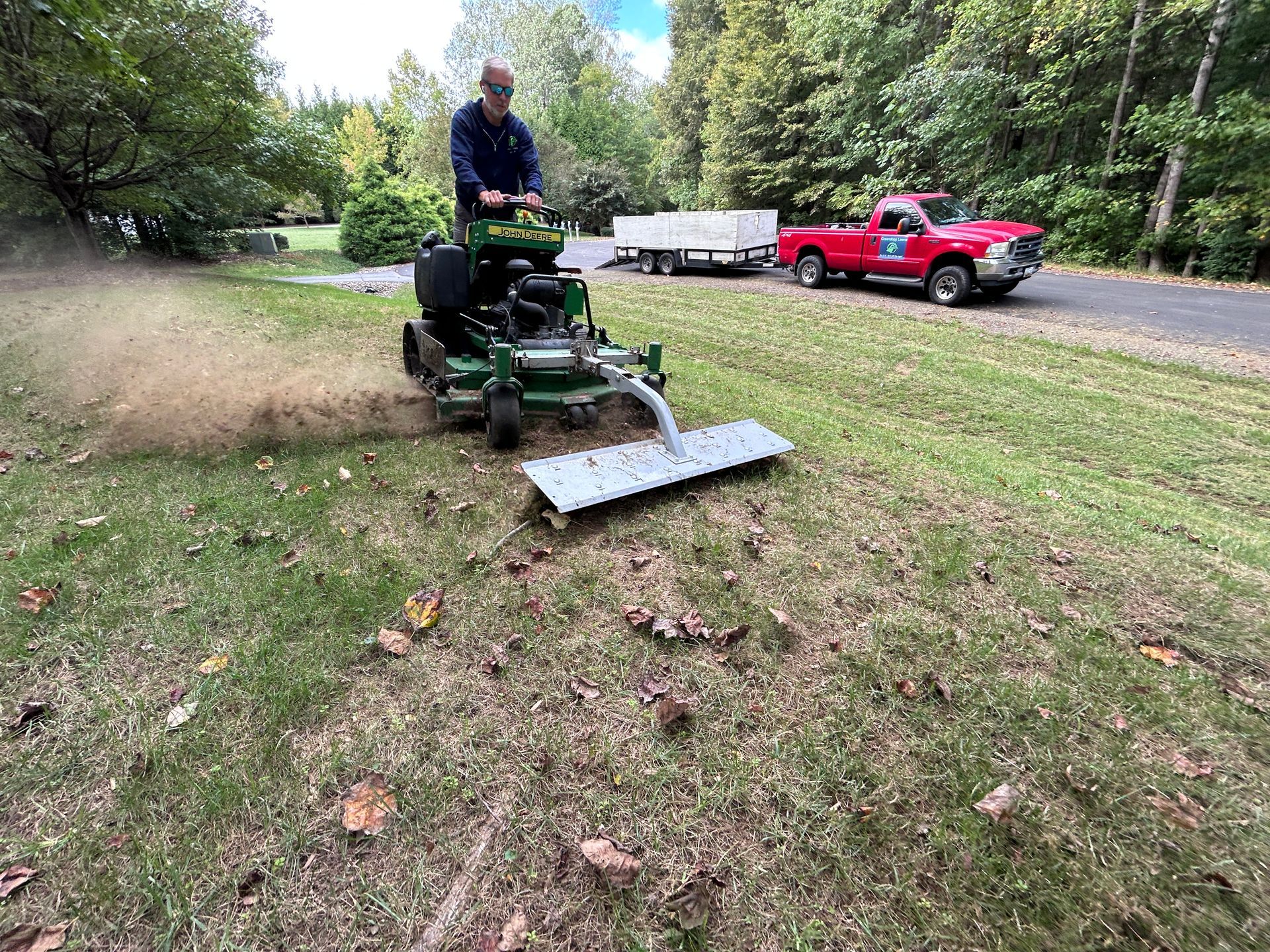 Man on a lawnmower blowing leaves. Red truck and trailer parked nearby.