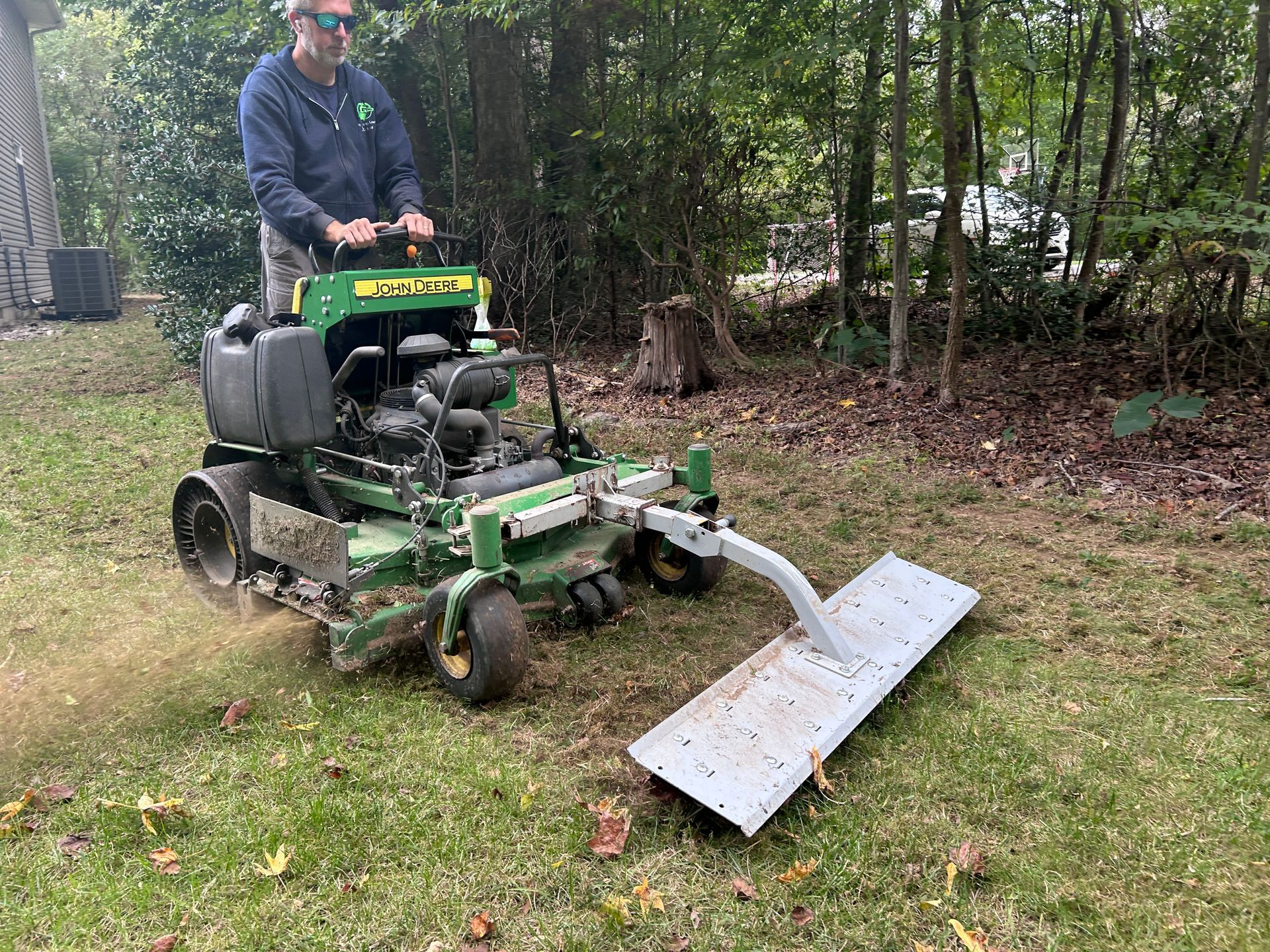 Man operating a John Deere lawnmower with a debris sweeper on grass in a yard.