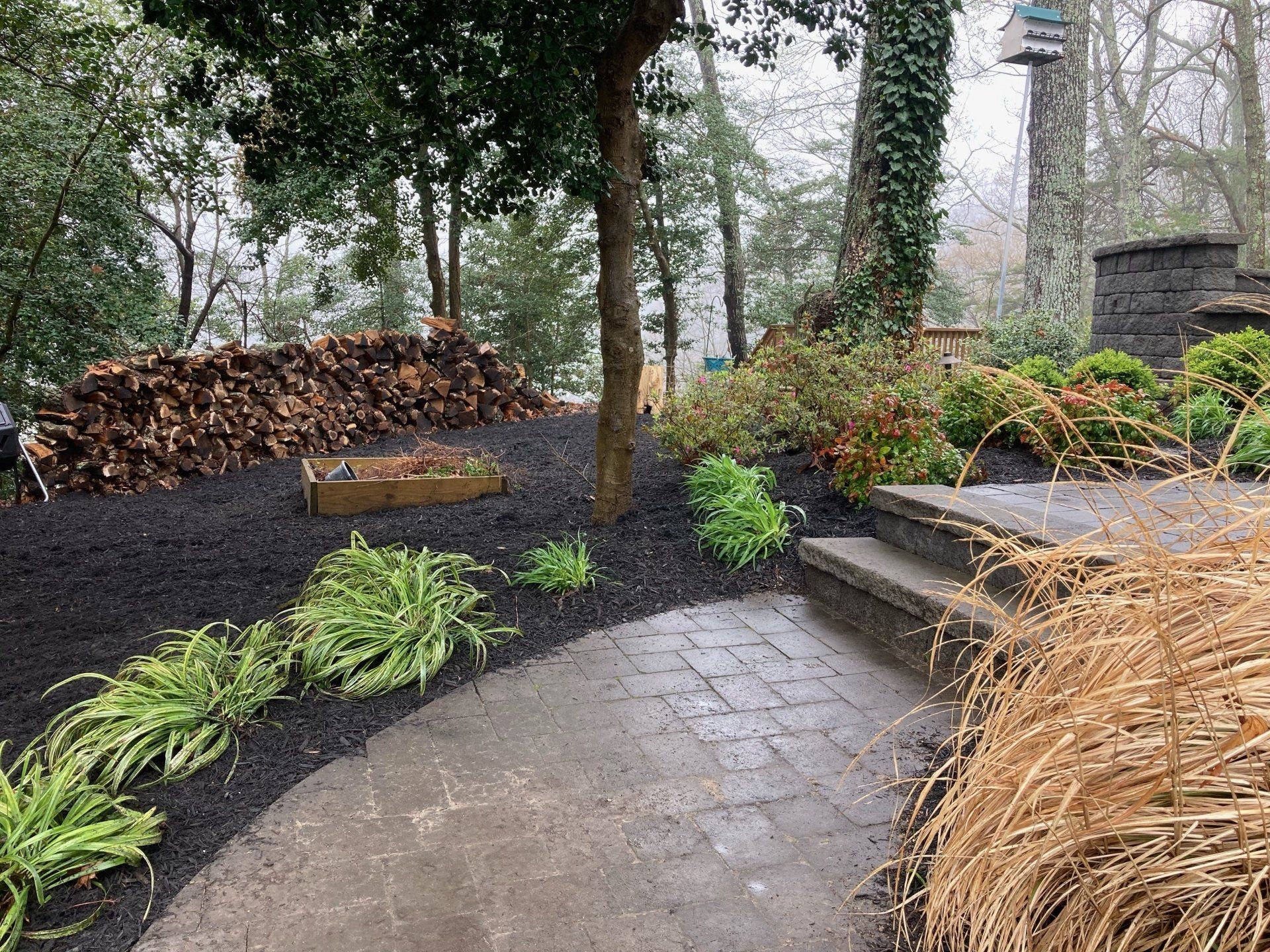 A brick walkway leading to a pile of wood in a garden.