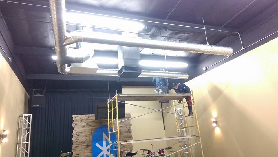 A man is standing on a scaffolding in a room with pipes hanging from the ceiling.