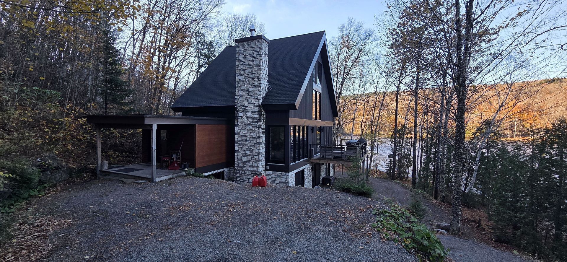 Un chalet moderne en forme de A avec une cheminée en pierre se dresse au bord d'un lac, en pleine forêt, en automne.