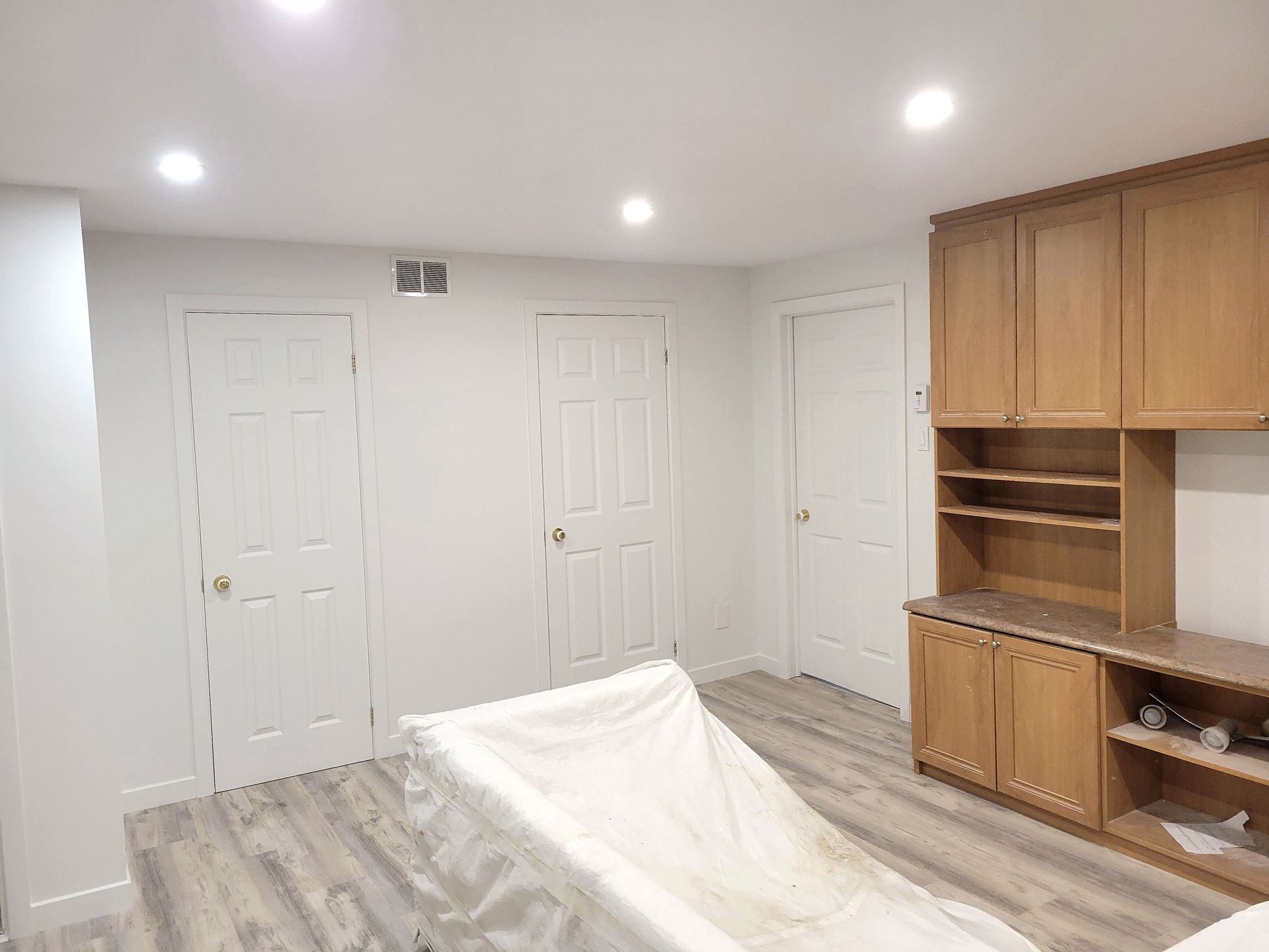 Une pièce avec un sol clair imitation bois, trois portes blanches, un éclairage encastré et une étagère en bois intégrée.