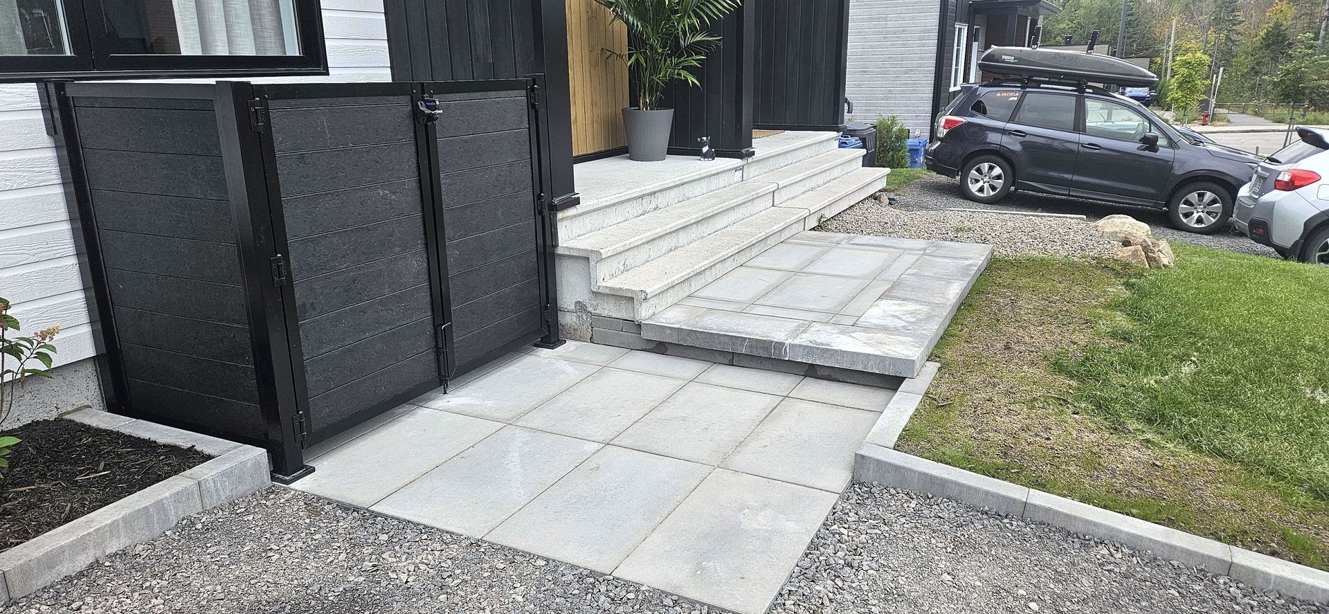 Un hangar noir se trouve à côté d'une allée en pierre gris clair menant aux marches d'un bâtiment, avec une cour en gravier et une voiture à proximité.
