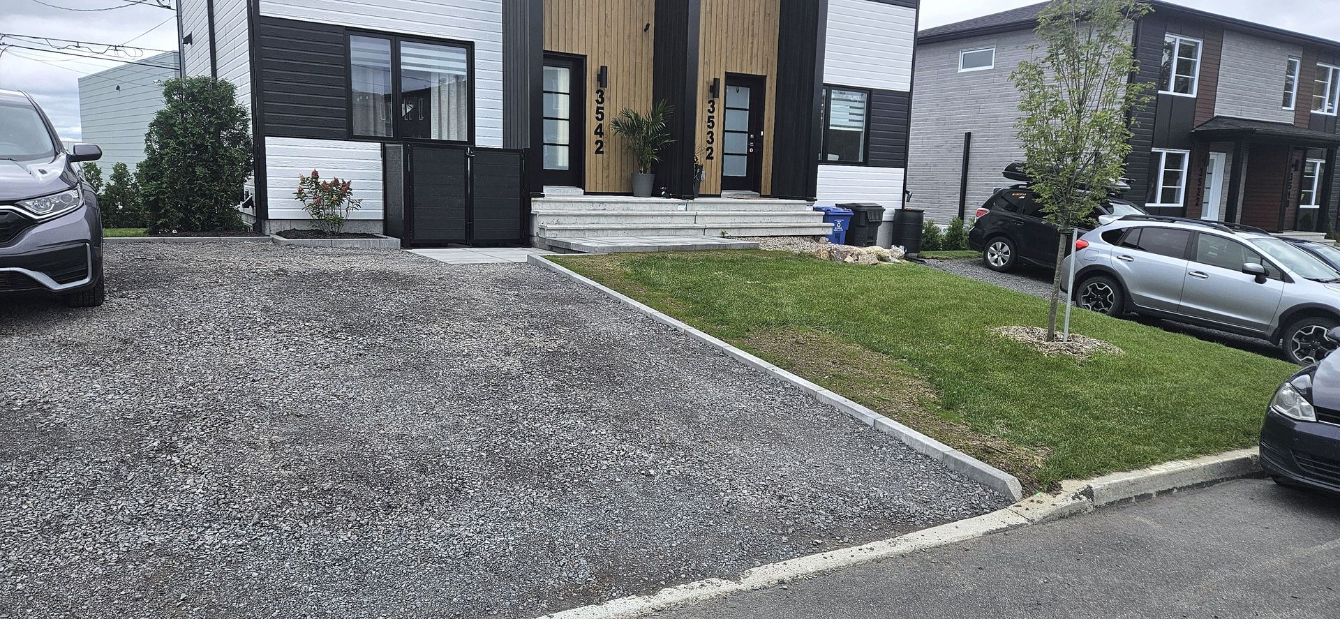 Un duplex moderne avec une allée en gravier gris, un petit carré de pelouse et des voitures garées devant.