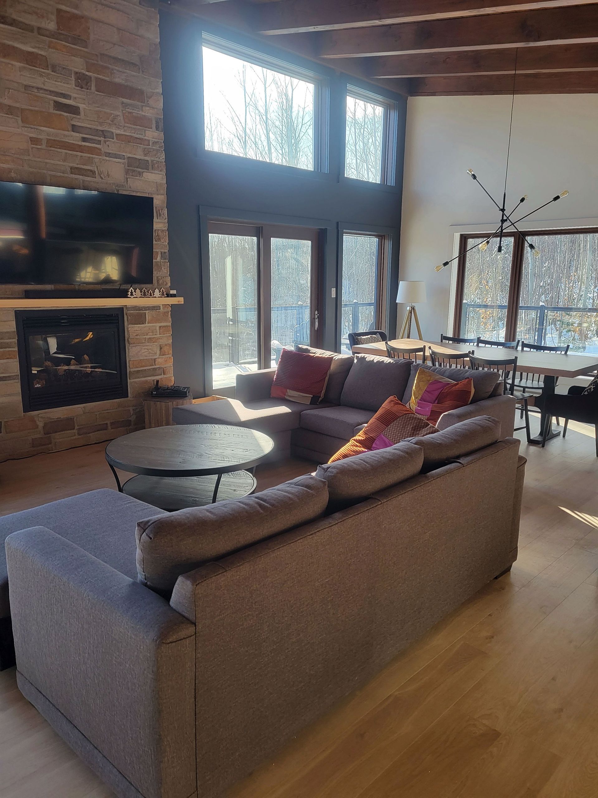 Un salon spacieux avec un grand canapé d'angle gris, une cheminée en pierre et de hautes fenêtres donnant sur une zone boisée.