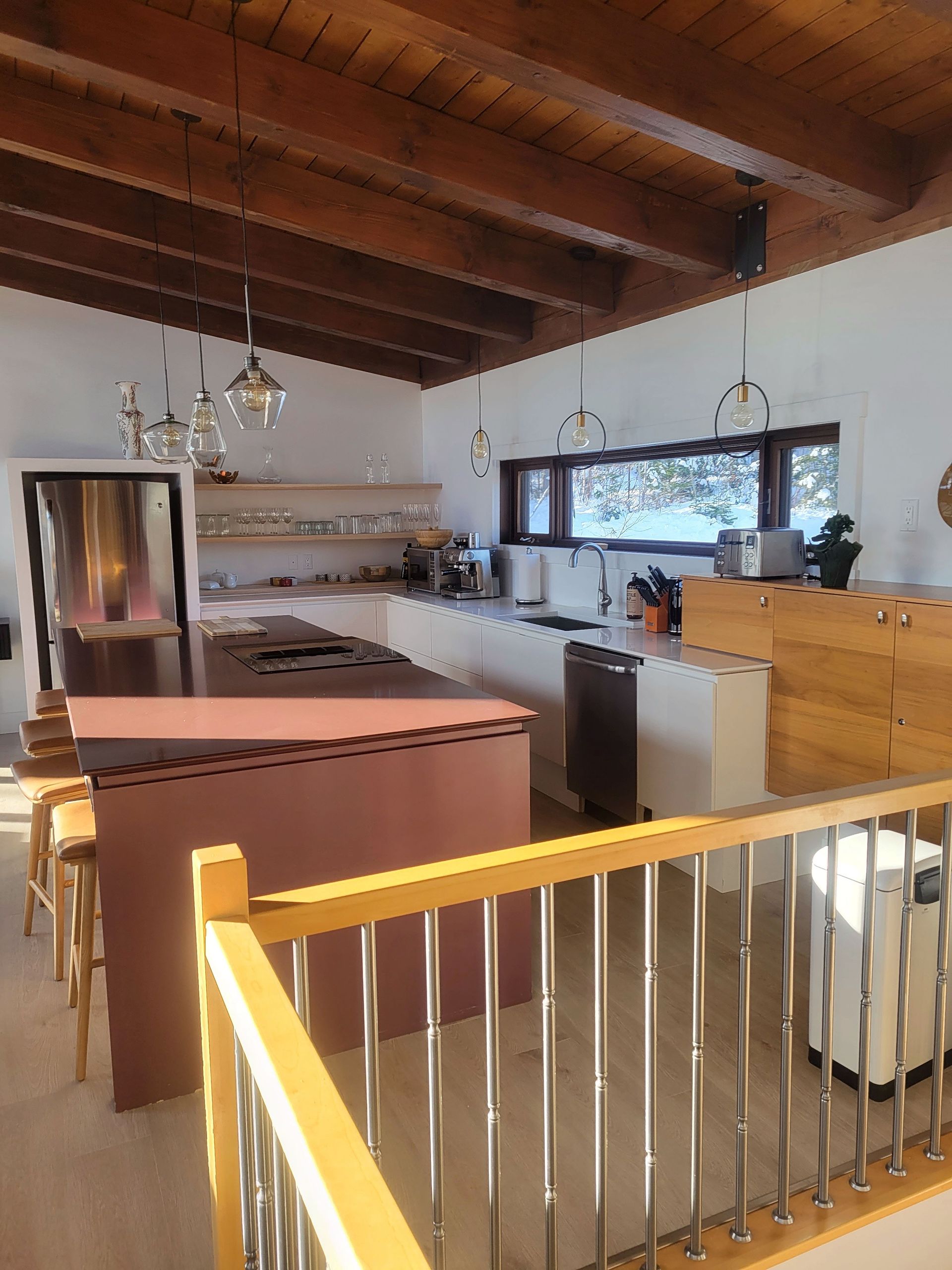 Une cuisine moderne avec un grand îlot central, des tabourets de bar, des poutres apparentes au plafond et une vue par la fenêtre, vue depuis un balcon avec rambarde.