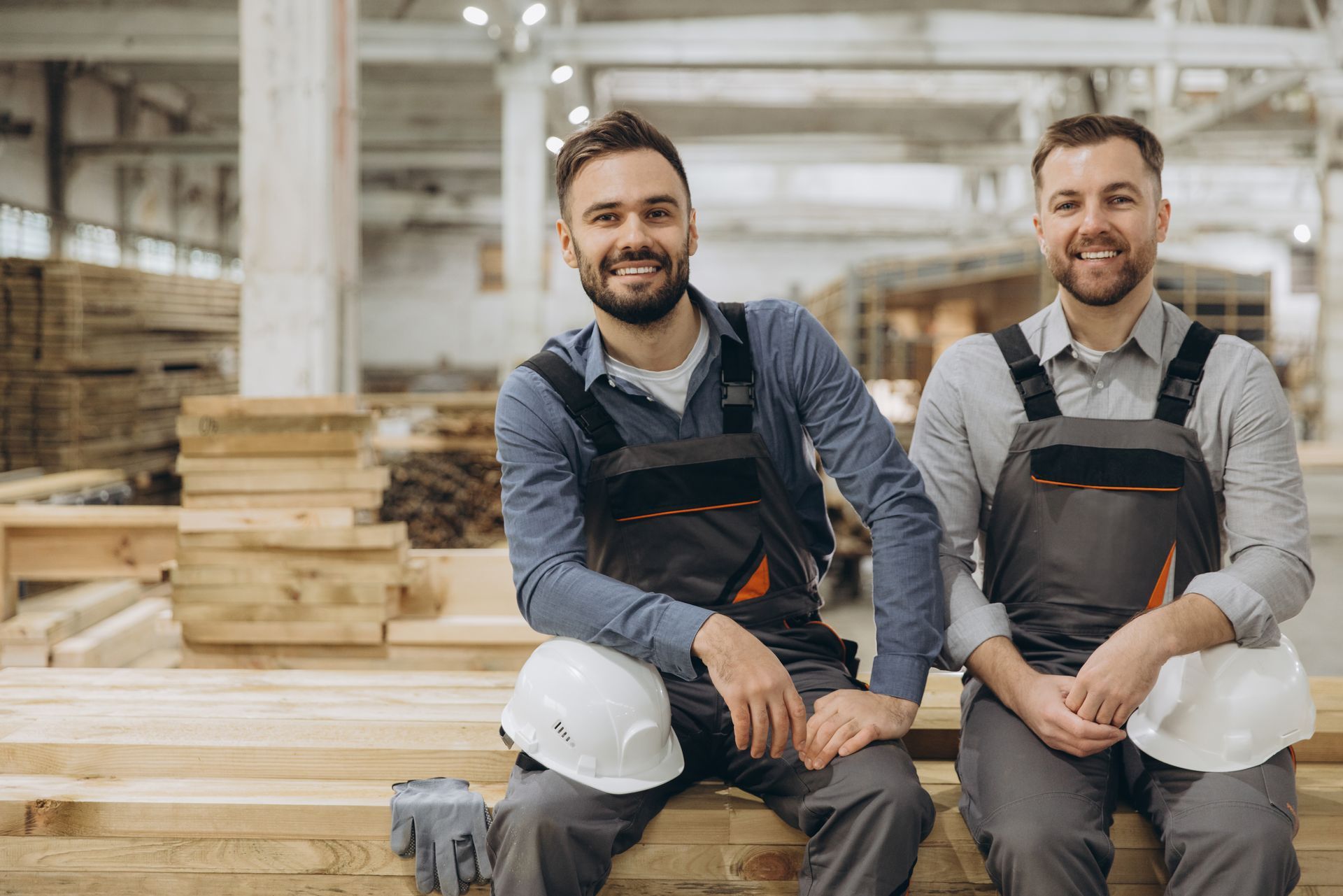 Deux ouvriers souriants, vêtus de vêtements de travail gris et de casques de chantier, sont assis sur des poutres en bois empilées dans un entrepôt lumineux.