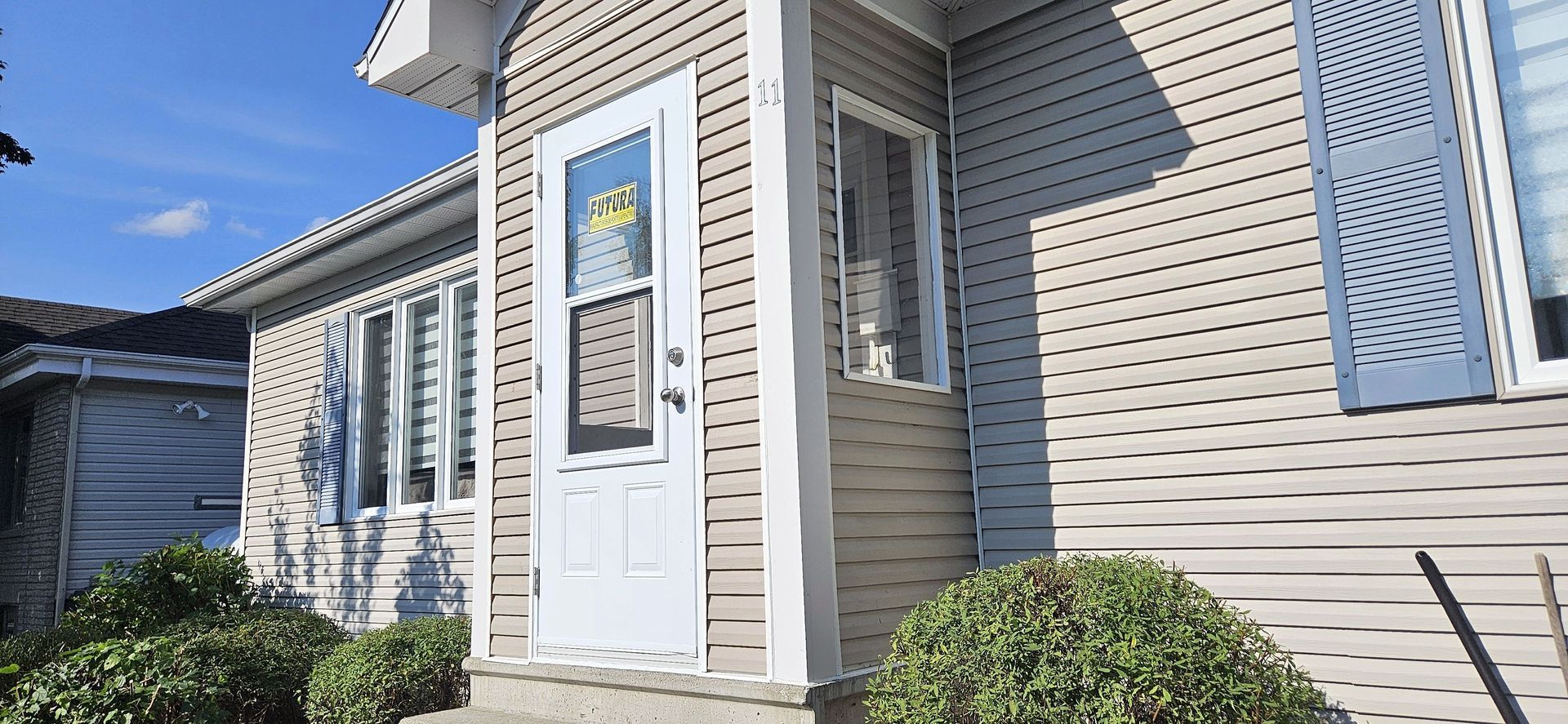 Vue de l'entrée d'une maison avec un bardage horizontal beige, une porte d'entrée blanche et des buissons verts sur un ciel bleu clair.