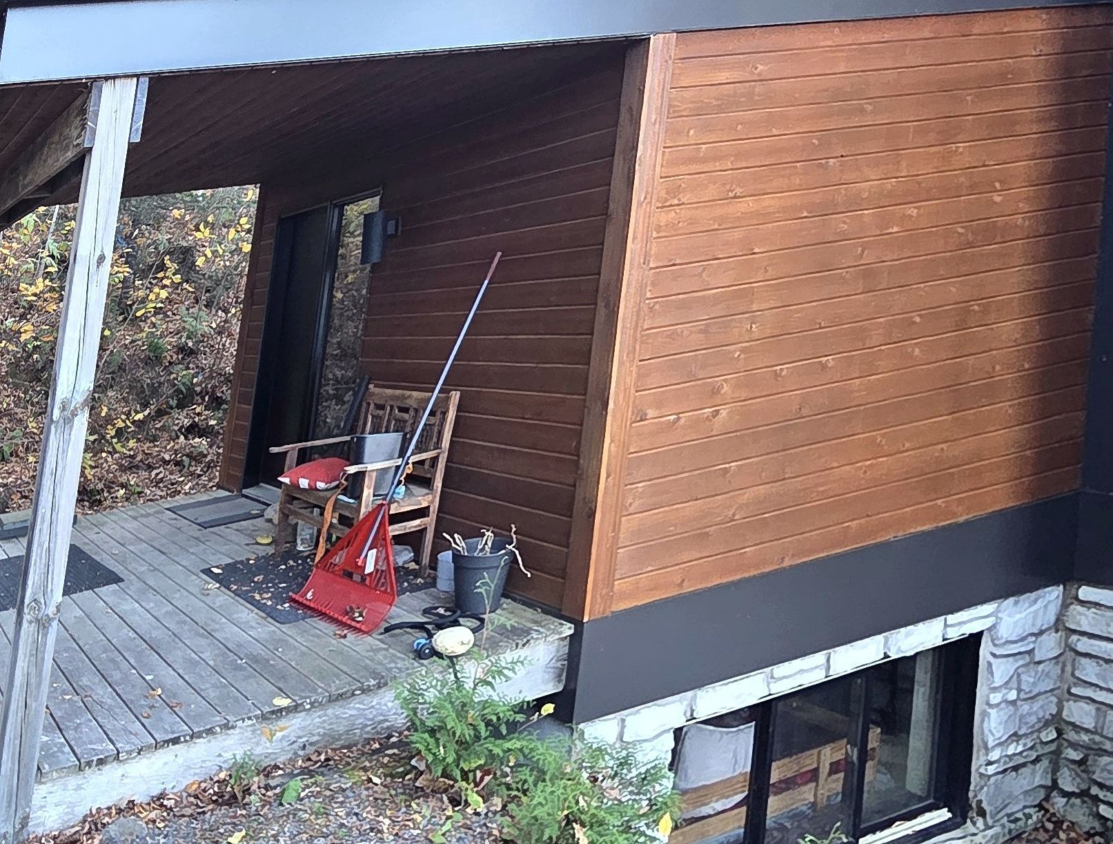 Mur extérieur en bois avec bardage horizontal au-dessus de fondations en pierre, comportant un porche avec une chaise et du matériel.