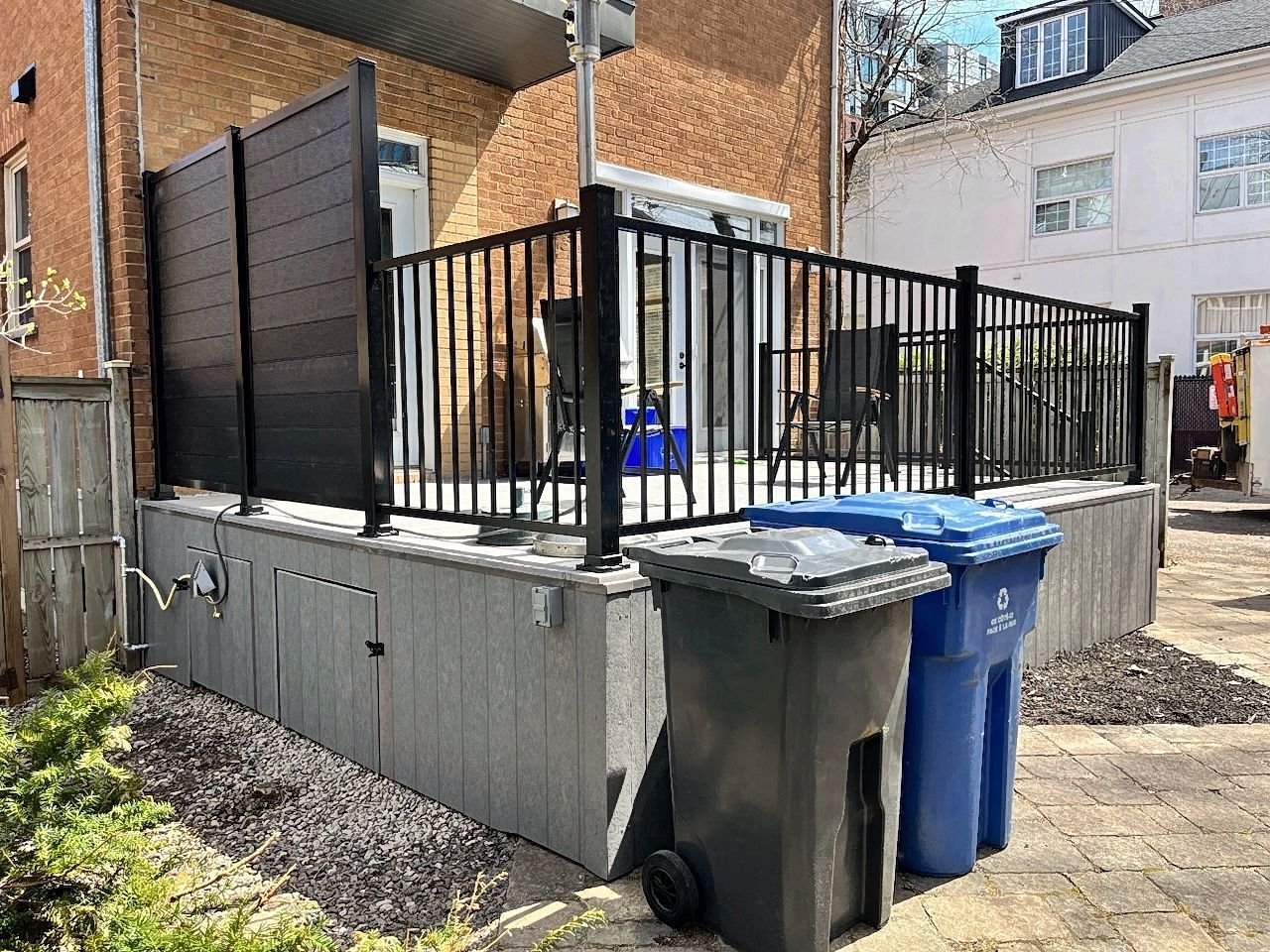 Dans une cour arrière, une terrasse surélevée grise, dotée d'une rambarde noire et d'un écran d'intimité, laisse apparaître des poubelles au premier plan.