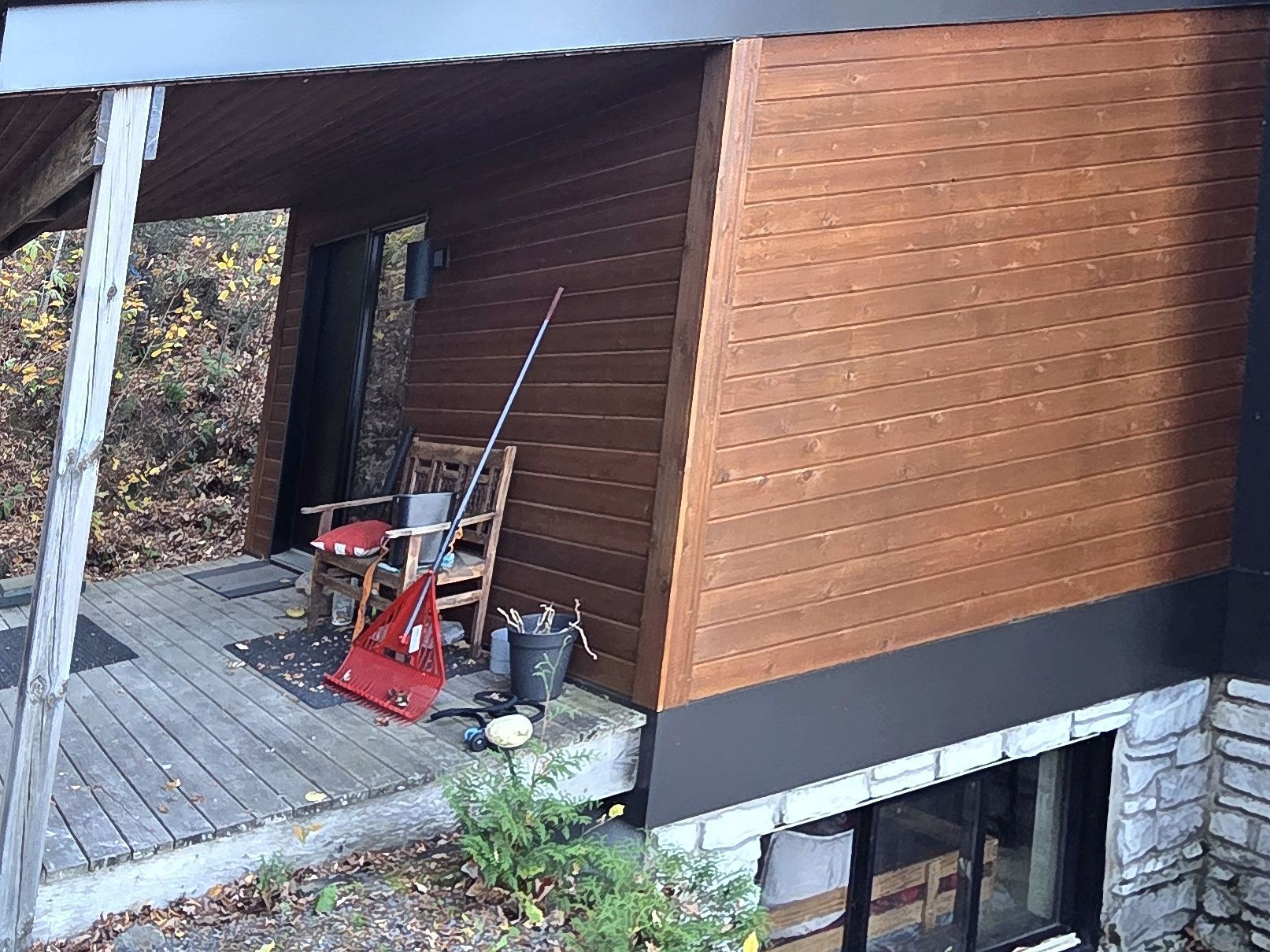 Une maison en bois avec une terrasse aménagée avec une chaise et des outils éparpillés, adossée à un mur extérieur en pente et à des fondations en pierre.