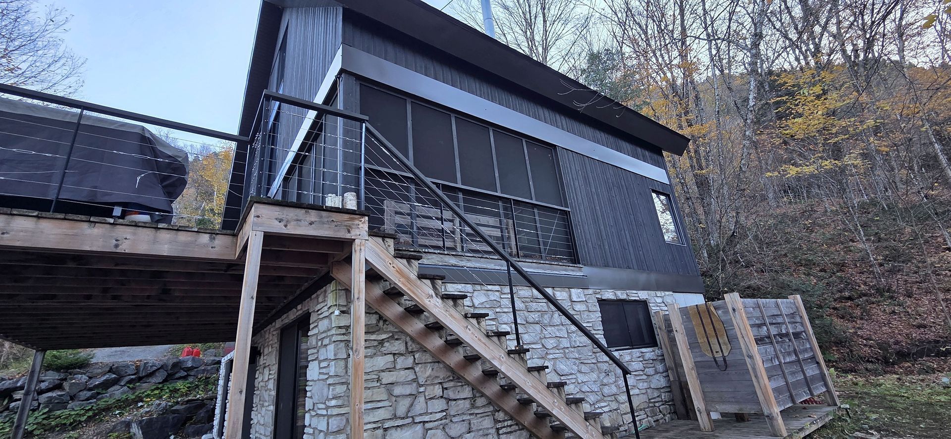 Un chalet moderne avec un bardage vertical gris foncé et un soubassement en pierre, doté d'une terrasse surélevée et d'un escalier en bois.