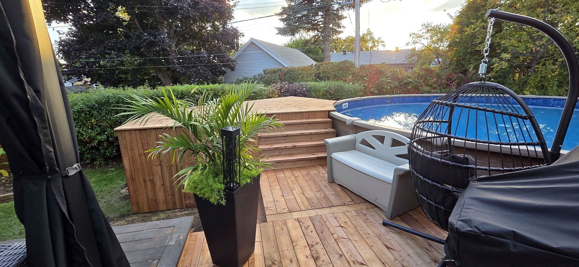 Terrasse arrière avec piscine surélevée en bois, escaliers à plusieurs niveaux, grande plante en pot, banc de rangement et fauteuil suspendu.