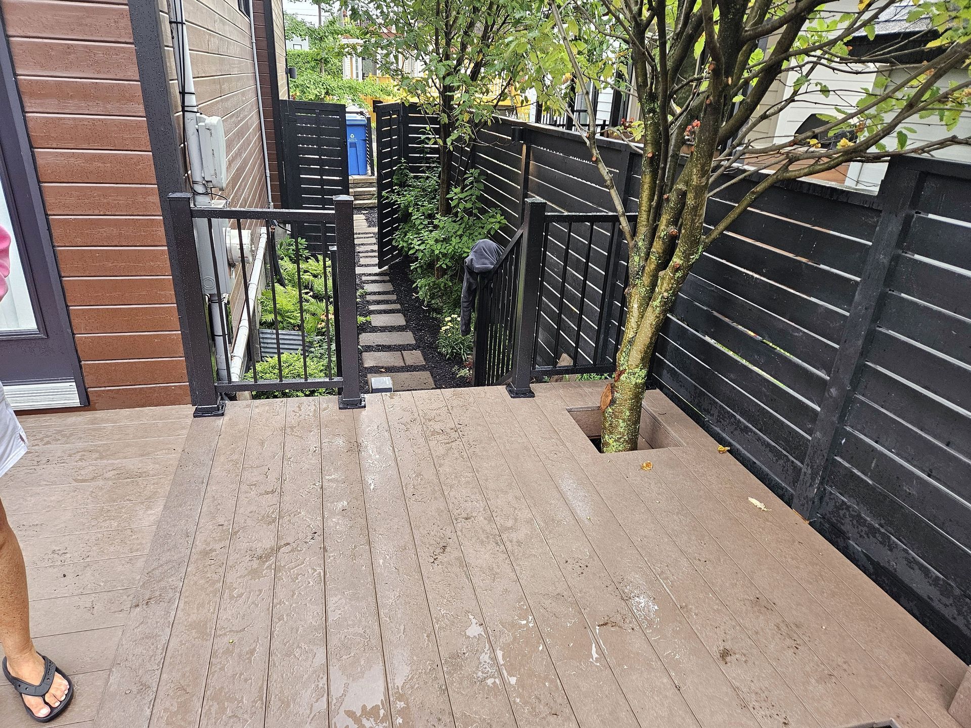 Une terrasse aux tons boisés présente un arbre qui pousse à travers une ouverture, avec une clôture noire, un portail et un chemin de pierre menant à un jardin.