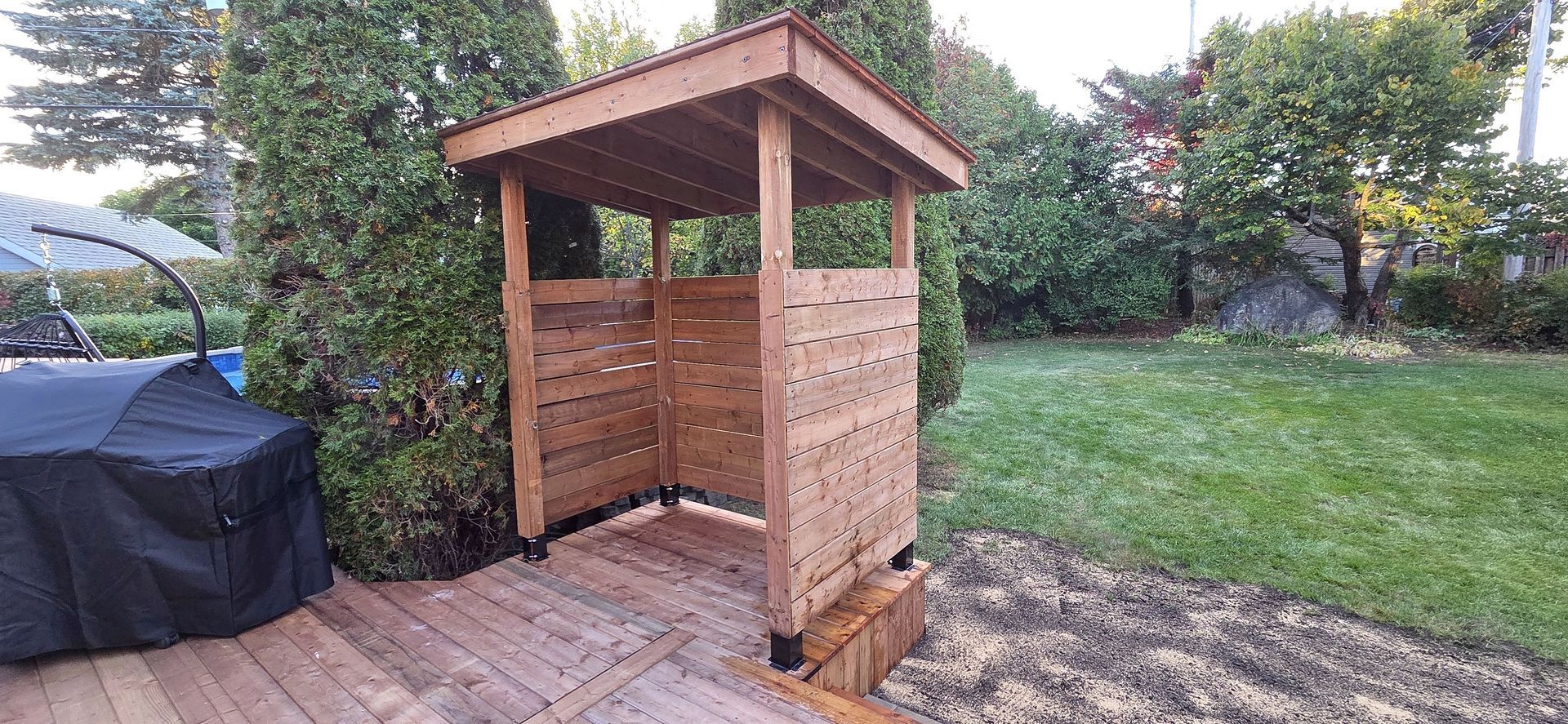 Un abri de jardin en bois, ouvert sur le devant, se dresse sur une terrasse dans une cour arrière, entouré d'arbres et de verdure.