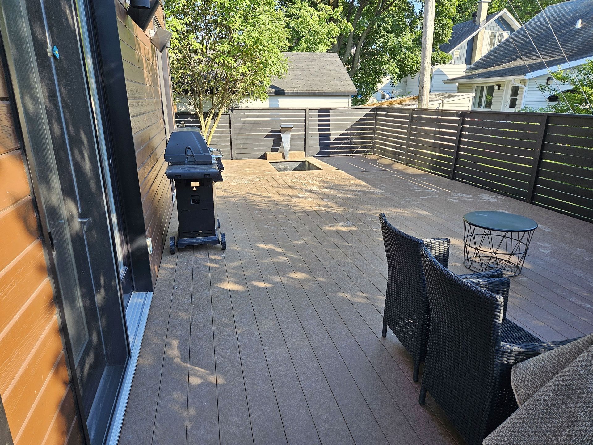 Une terrasse arrière avec un revêtement de sol composite marron, un barbecue noir, deux chaises noires et une table d'appoint en métal, le tout entouré d'une clôture.