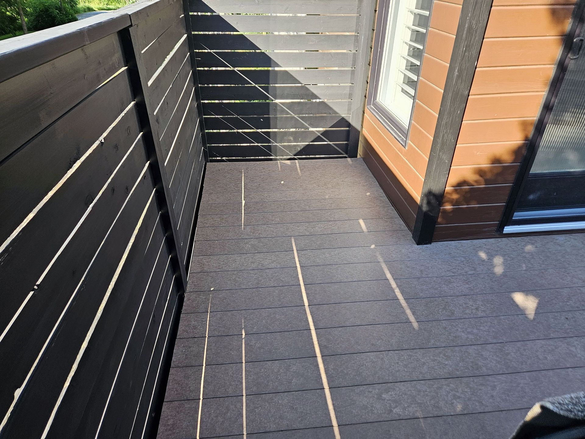 Une petite terrasse extérieure avec un revêtement de sol modulaire gris foncé et des panneaux d'intimité à lattes noires, adjacente à un mur lambrissé.
