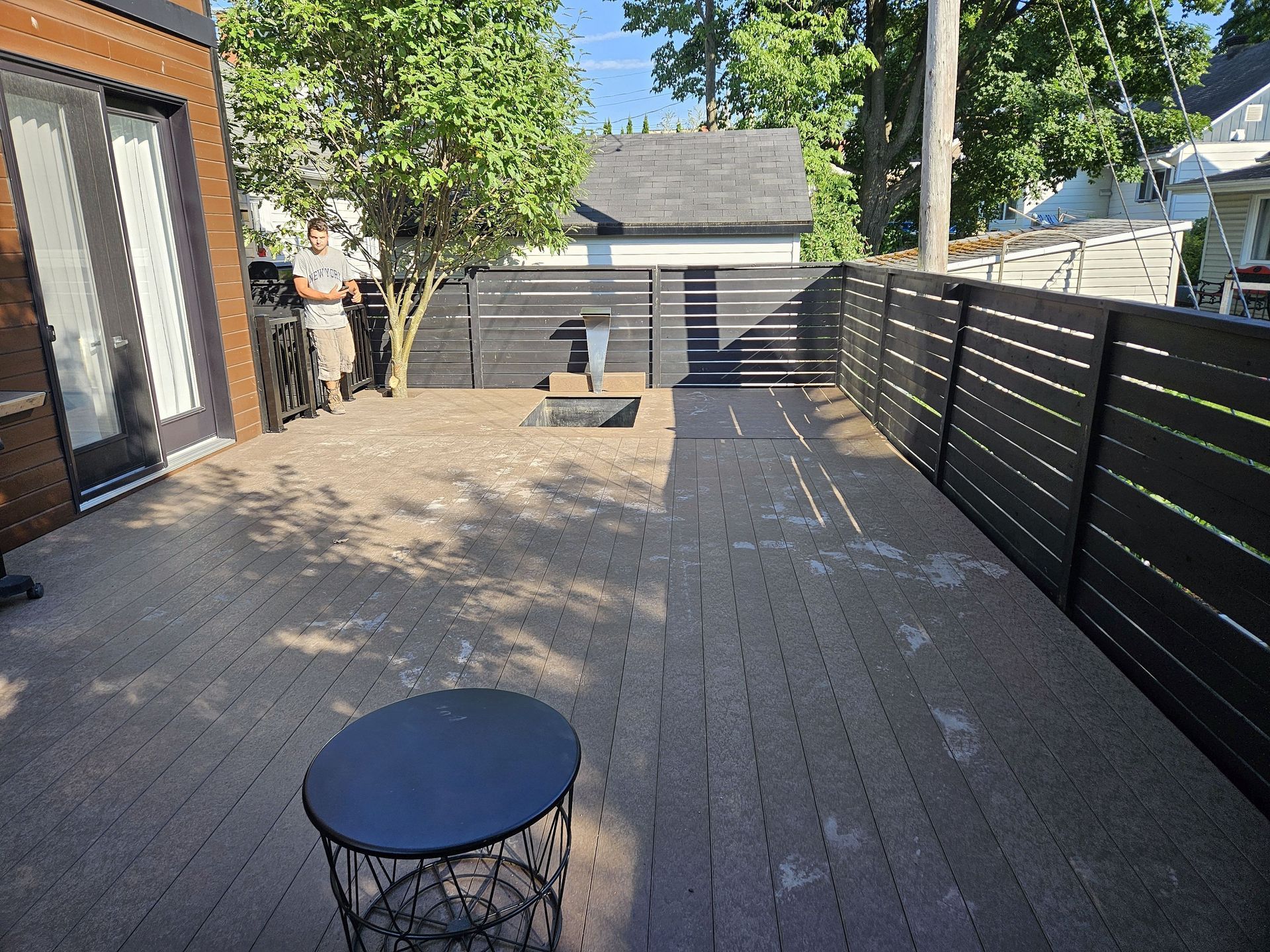 Une personne se tient debout sur une terrasse en composite marron, entourée d'une clôture à lattes noires pour préserver l'intimité, d'une petite table ronde et d'un arbre dans le jardin.