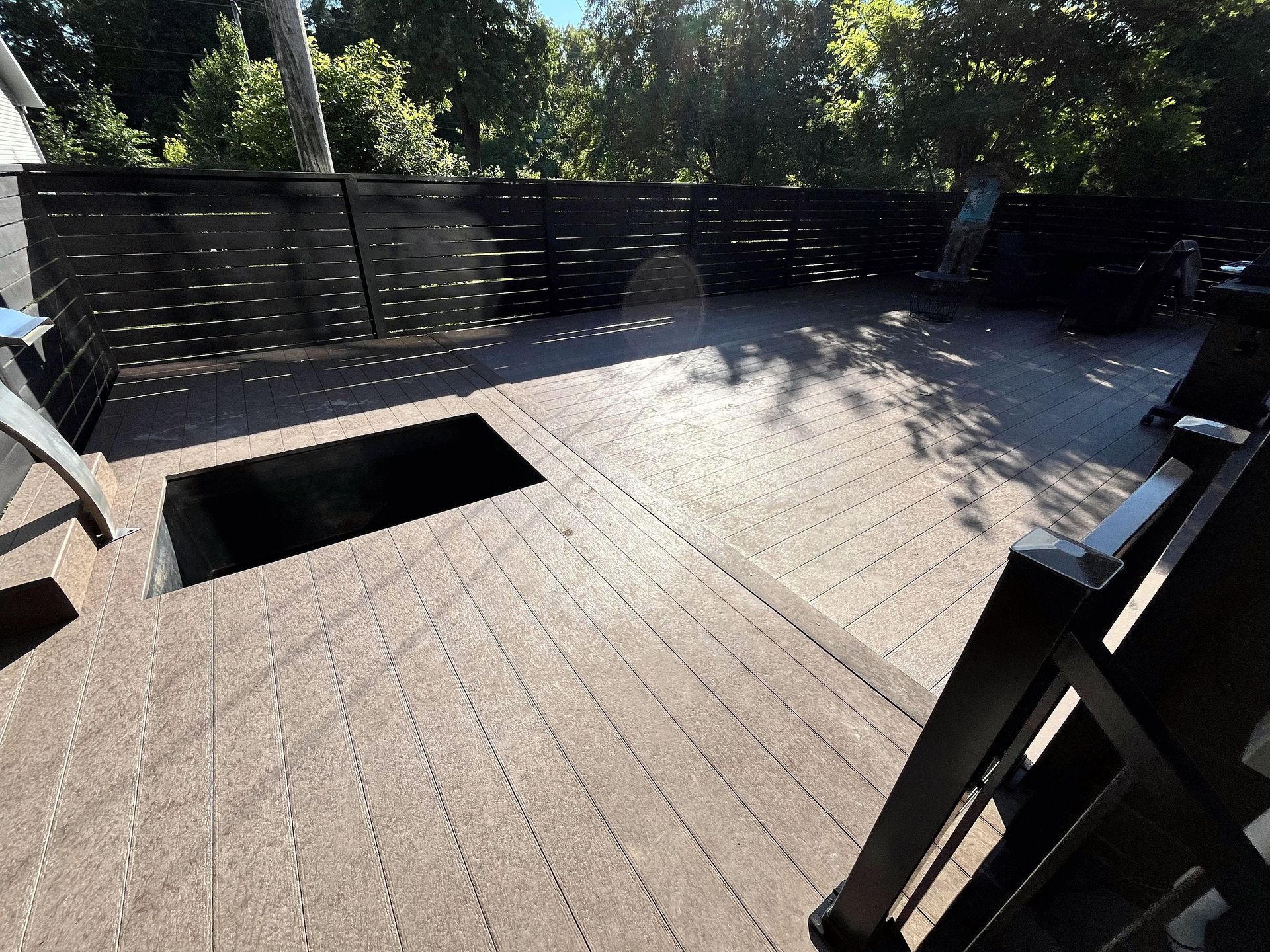 Une terrasse en bois avec une ouverture rectangulaire intégrée, entourée d'une clôture à claire-voie noire, baignée de soleil.