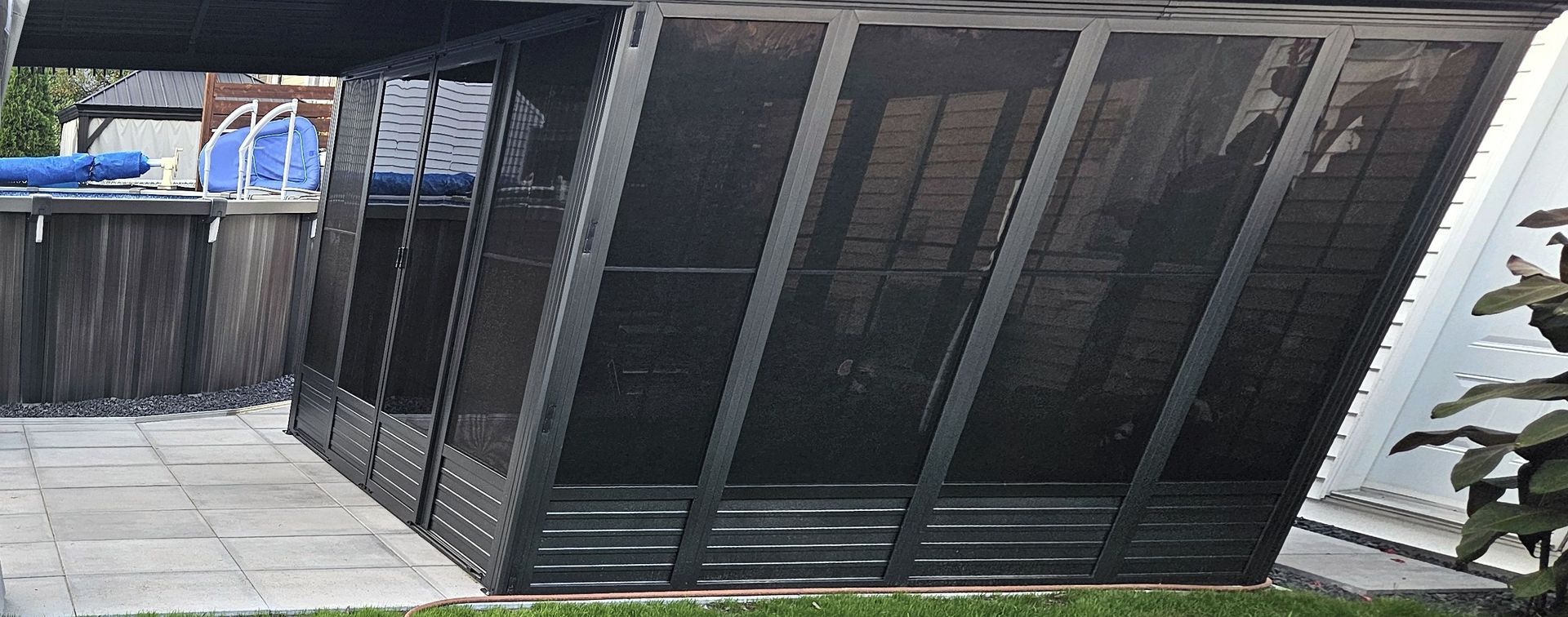 Une structure de type gazebo en métal noir, de forme angulaire, avec des parois grillagées, installée sur une terrasse en pierre à côté d'une piscine hors sol.