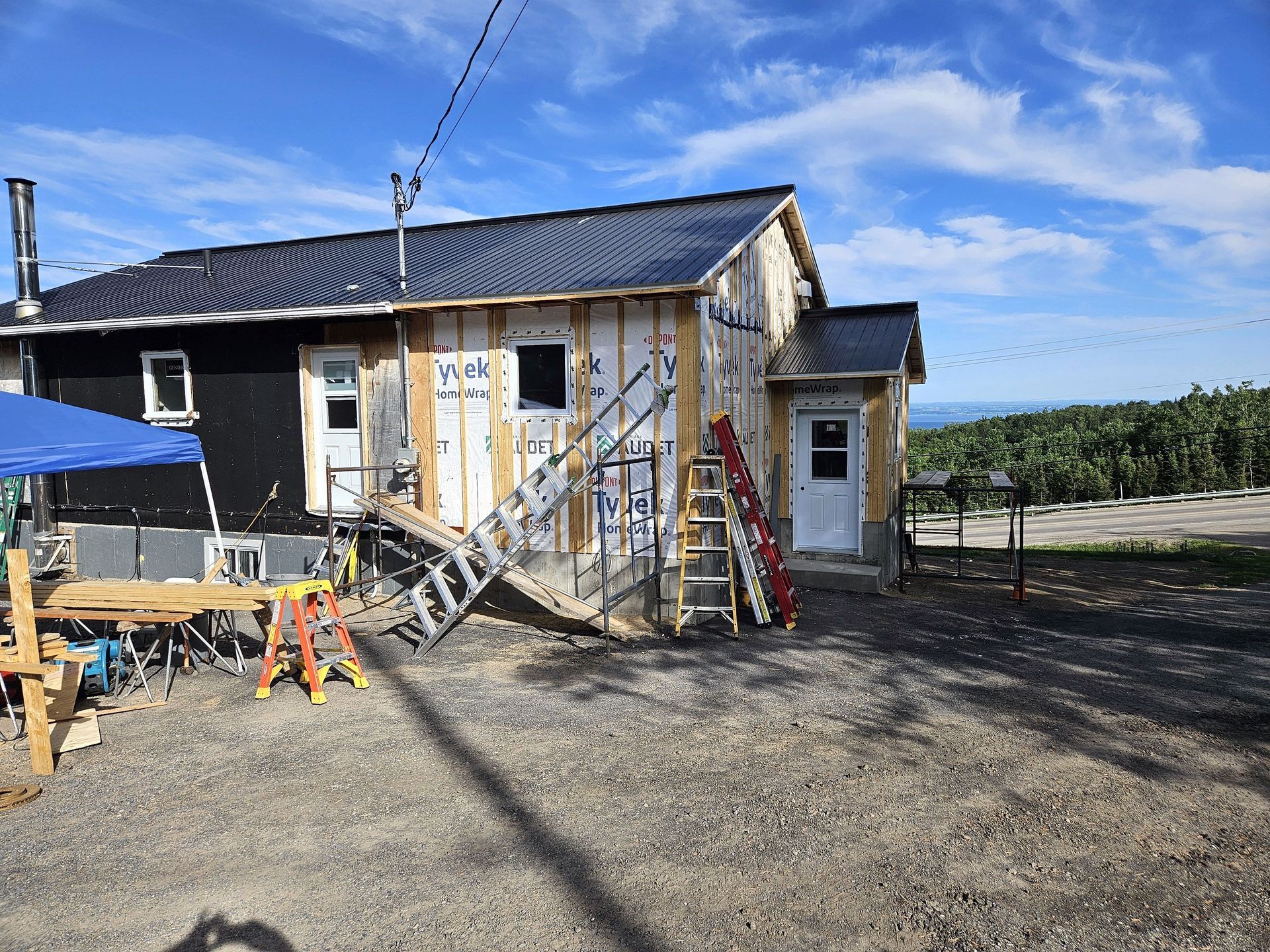 Une maison en rénovation, avec une charpente en bois apparente, des panneaux extérieurs noirs et un échafaudage sur un terrain gravillonné.