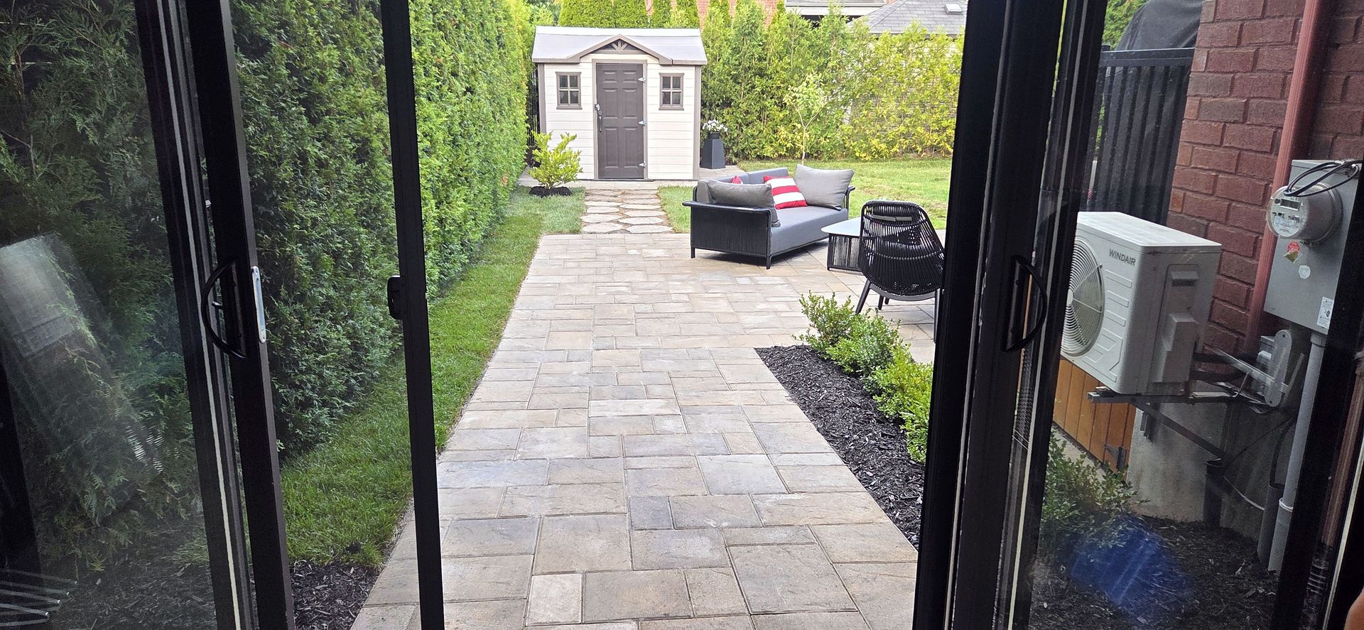 Un chemin pavé mène à un abri de jardin dans une cour arrière bordée de haies, agrémentée d'un canapé de patio et d'un climatiseur extérieur.