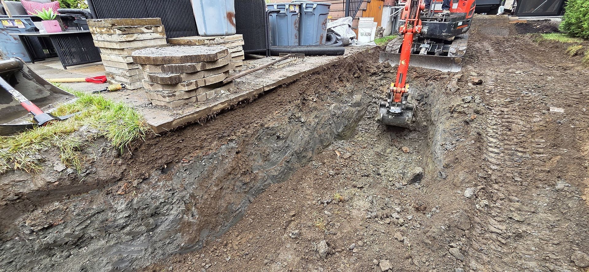 Une mini-pelle orange vif creuse une tranchée dans une cour résidentielle boueuse et encombrée de travaux.