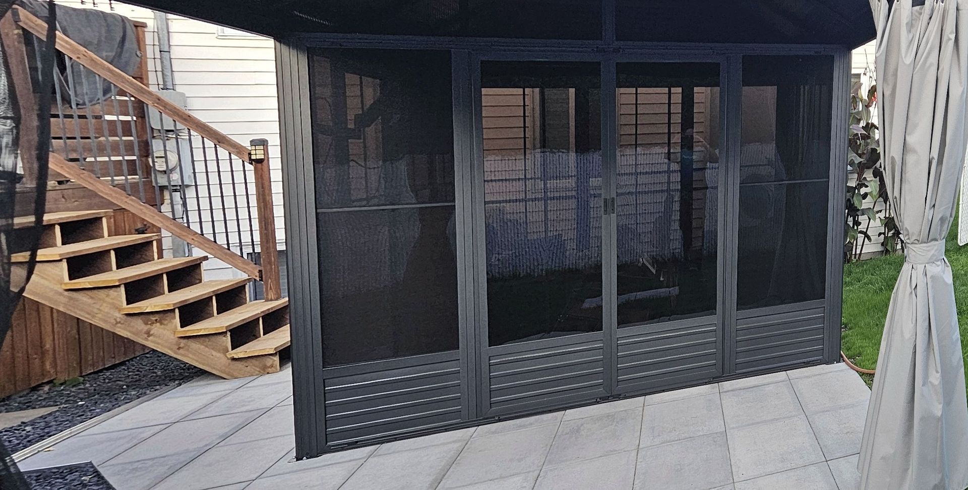 Un kiosque à la structure sombre et aux parois en filet se dresse sur une terrasse, à côté d'un escalier en bois menant à une terrasse en bois.