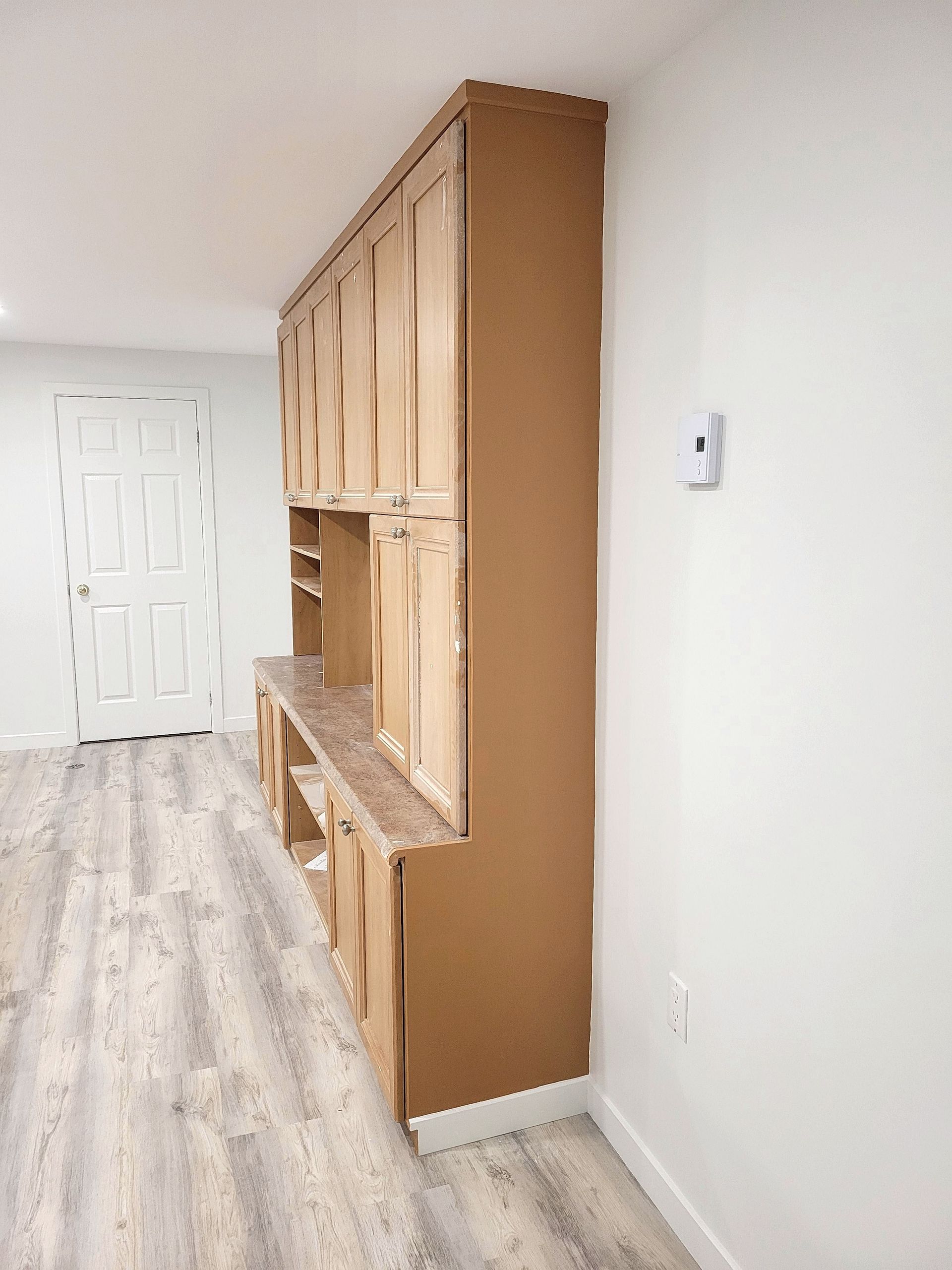 Dans une pièce au sol clair imitant le bois, un meuble encastré en bois avec un plan de travail est adossé à un mur blanc.