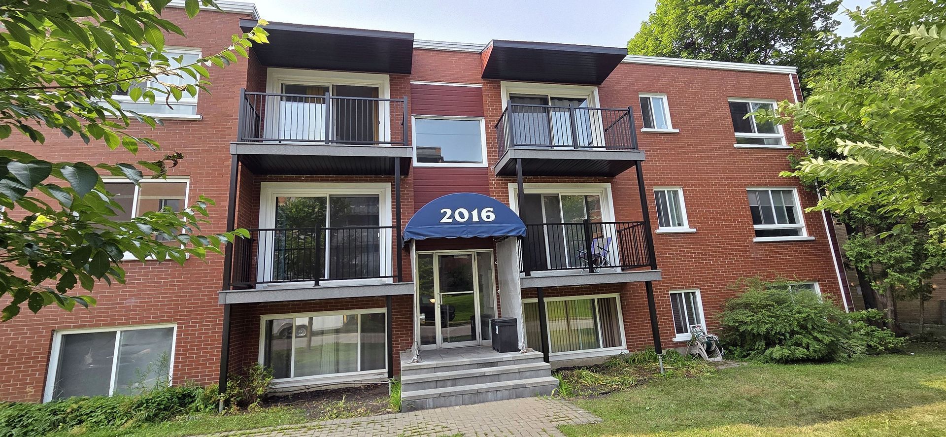 Un immeuble d'appartements de trois étages en briques rouges, avec des balcons noirs et un auvent bleu au-dessus de l'entrée, portant la mention 2016.
