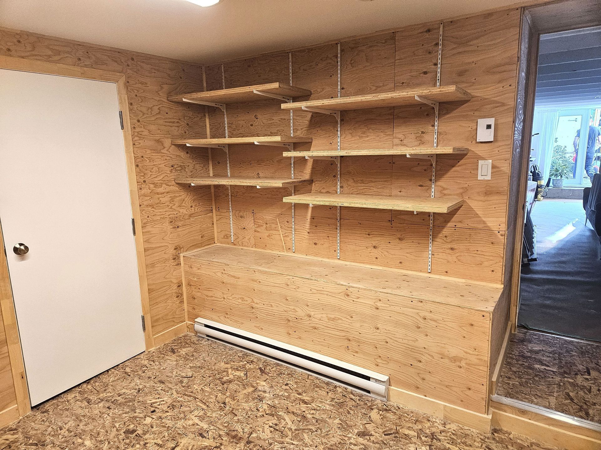 Une pièce aux murs en panneaux de particules de couleur claire, avec des étagères ouvertes sur rails métalliques au-dessus d'un comptoir et d'un radiateur intégrés.