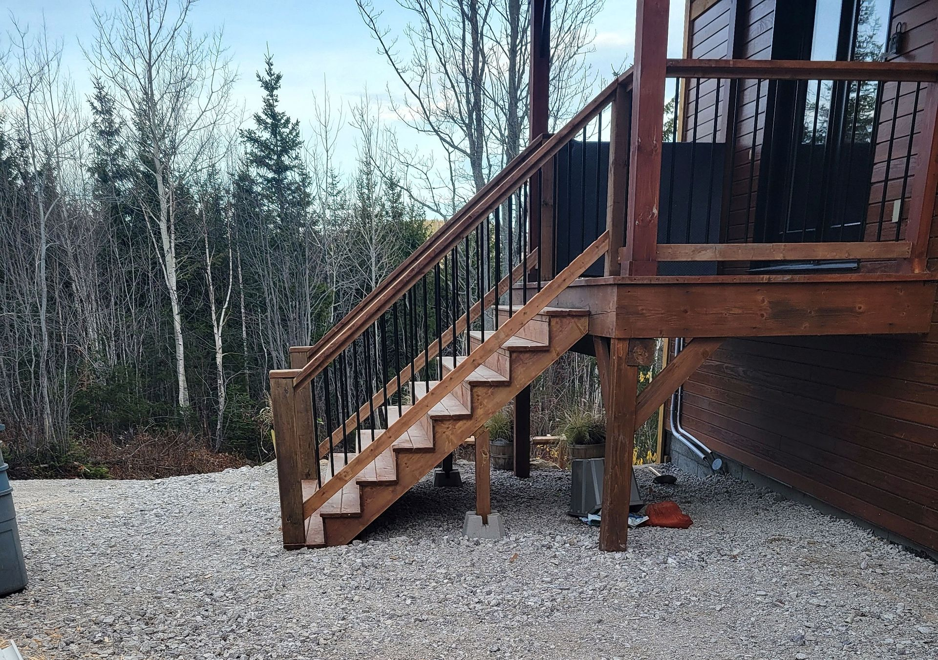 Un escalier en bois avec des rampes en métal noir mène à la terrasse d'un chalet en rondins entouré d'arbres sur un terrain gravillonné.