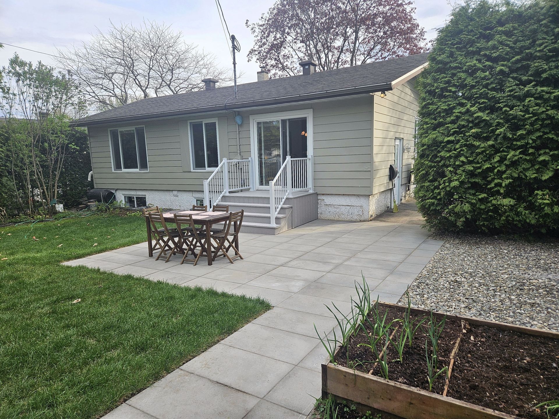 Une terrasse avec une table et des chaises installées devant une maison de plain-pied aux couleurs claires, dotée de portes coulissantes en verre.