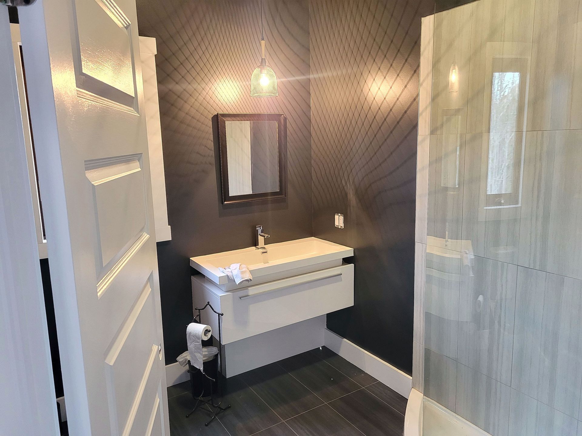 Une salle de bains moderne avec du papier peint à motifs foncés, un meuble-lavabo suspendu blanc, un petit miroir carré et une douche en verre.