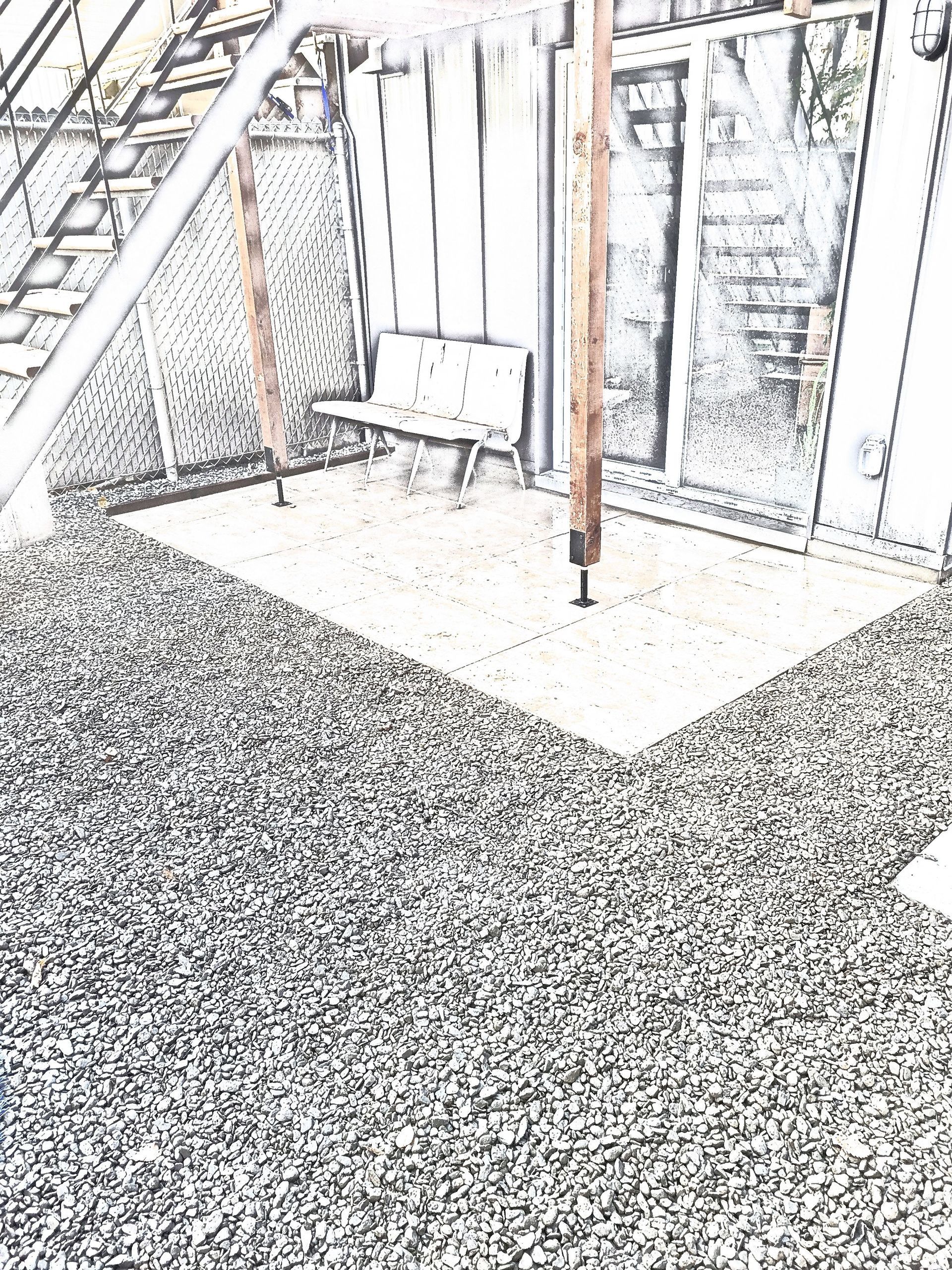Un patio aux tons gris avec un petit banc sous une structure en bois, adossé à un bâtiment avec un escalier et une porte vitrée.
