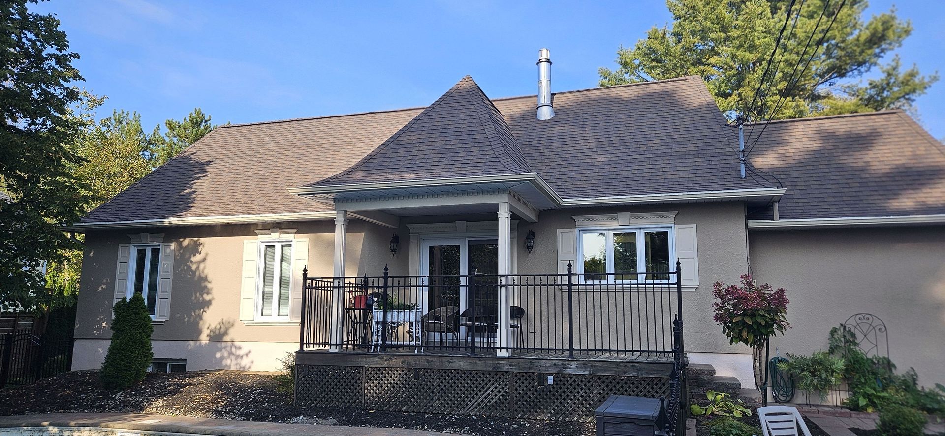 Une maison de plain-pied de couleur beige avec un toit en bardeaux, dotée d'un porche, d'une rambarde en métal et de fenêtres à volets blancs.