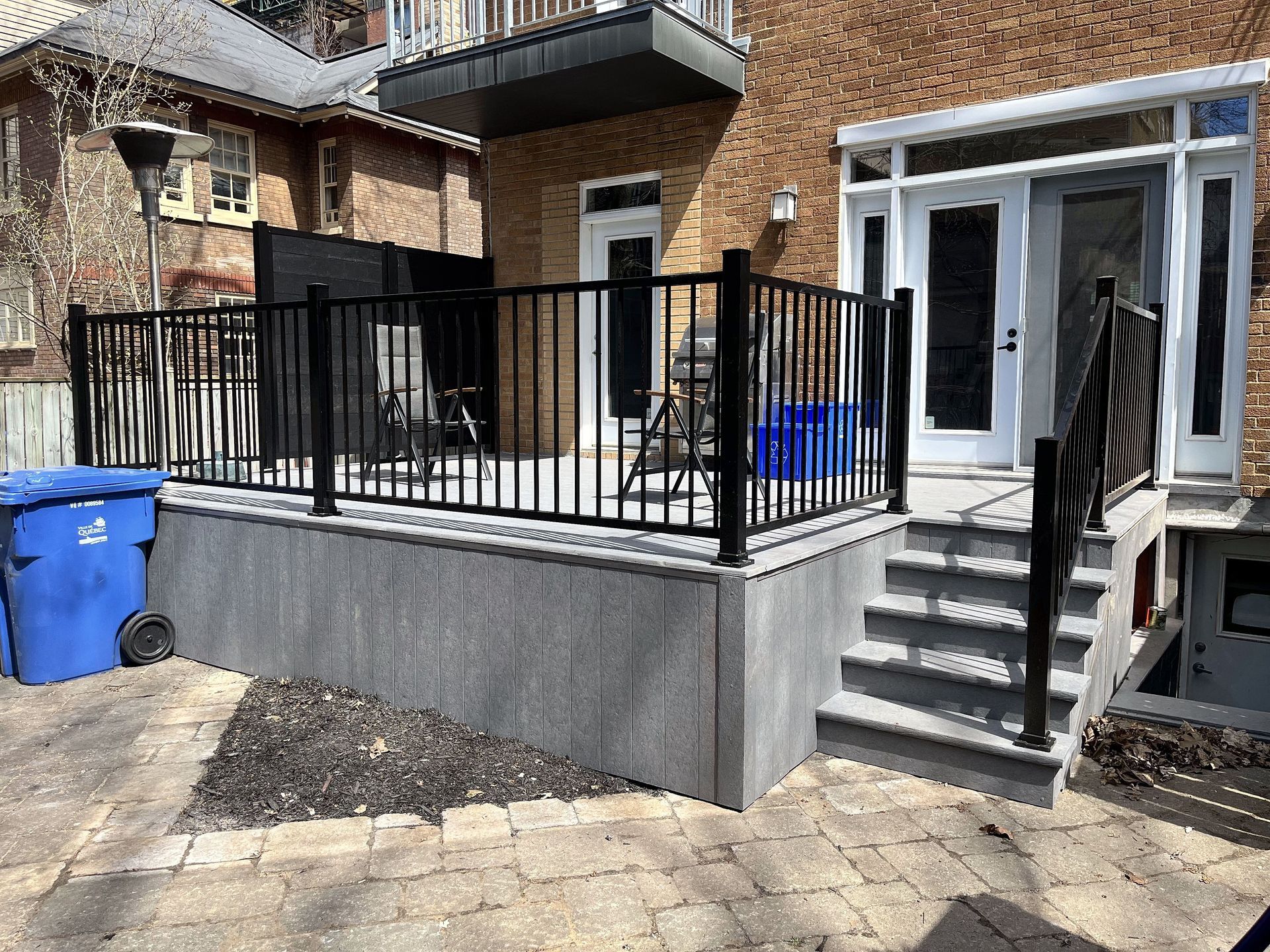 Une terrasse surélevée avec des rambardes en métal noir et des escaliers menant à une entrée arrière d'une maison en briques avec une poubelle bleue.
